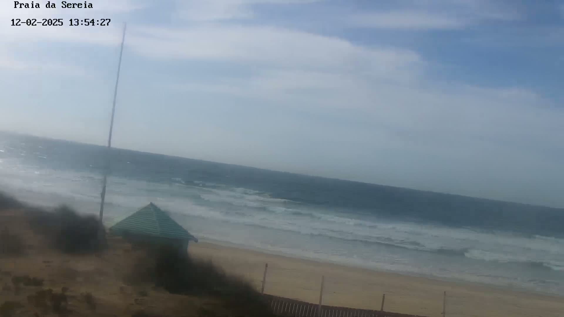 A sandy beach with a small green structure is shown, with a somewhat cloudy sky and moderately wavy ocean in the background.