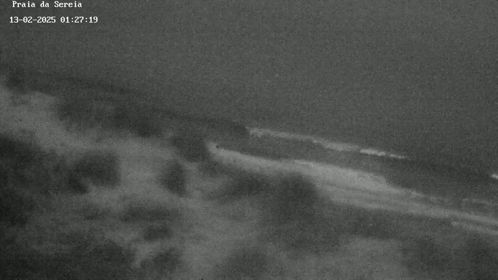 A nighttime, low-light image shows a dark beach with faint waves under an overcast sky.