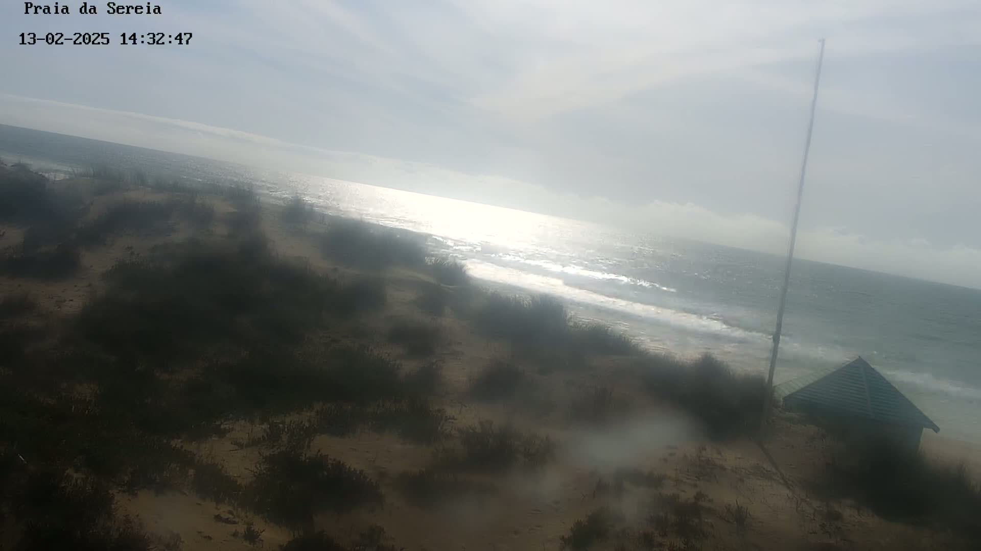A hazy, sunlit beach scene shows a sandy shore with low-lying vegetation, a small building, and a tall pole, overlooking a calm sea with gentle waves.