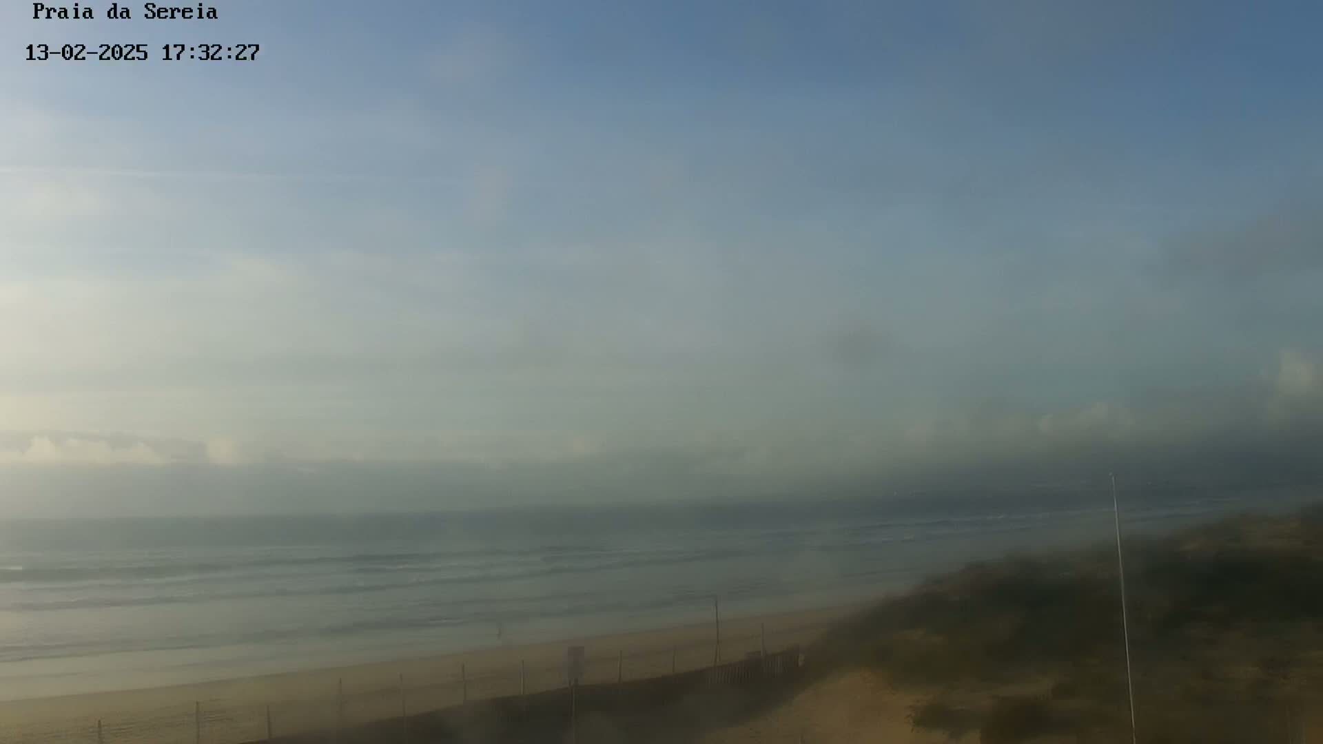 A hazy view shows a calm ocean with small waves lapping a sandy beach, partially obscured by fog.