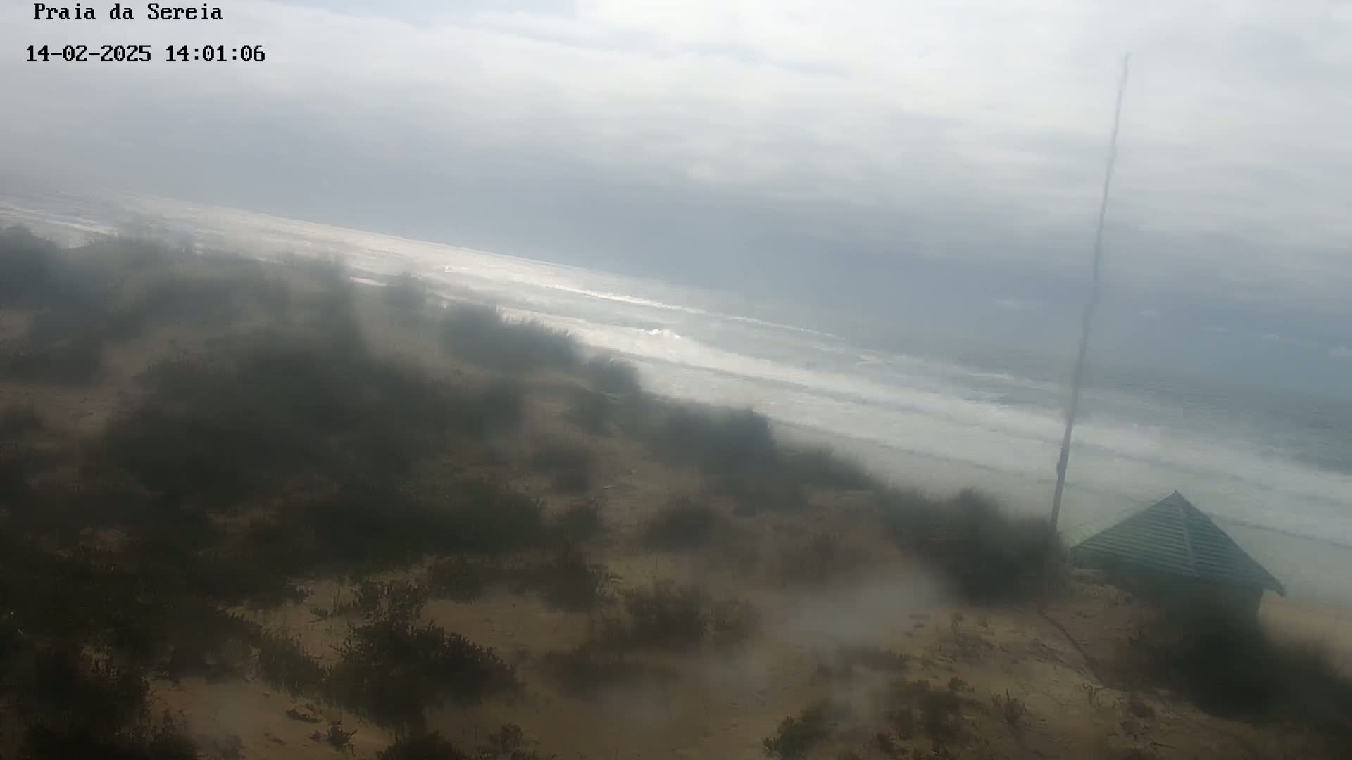 A hazy view shows a sandy beach with sparse vegetation, a small green building, and a calm ocean with small waves under an overcast sky.