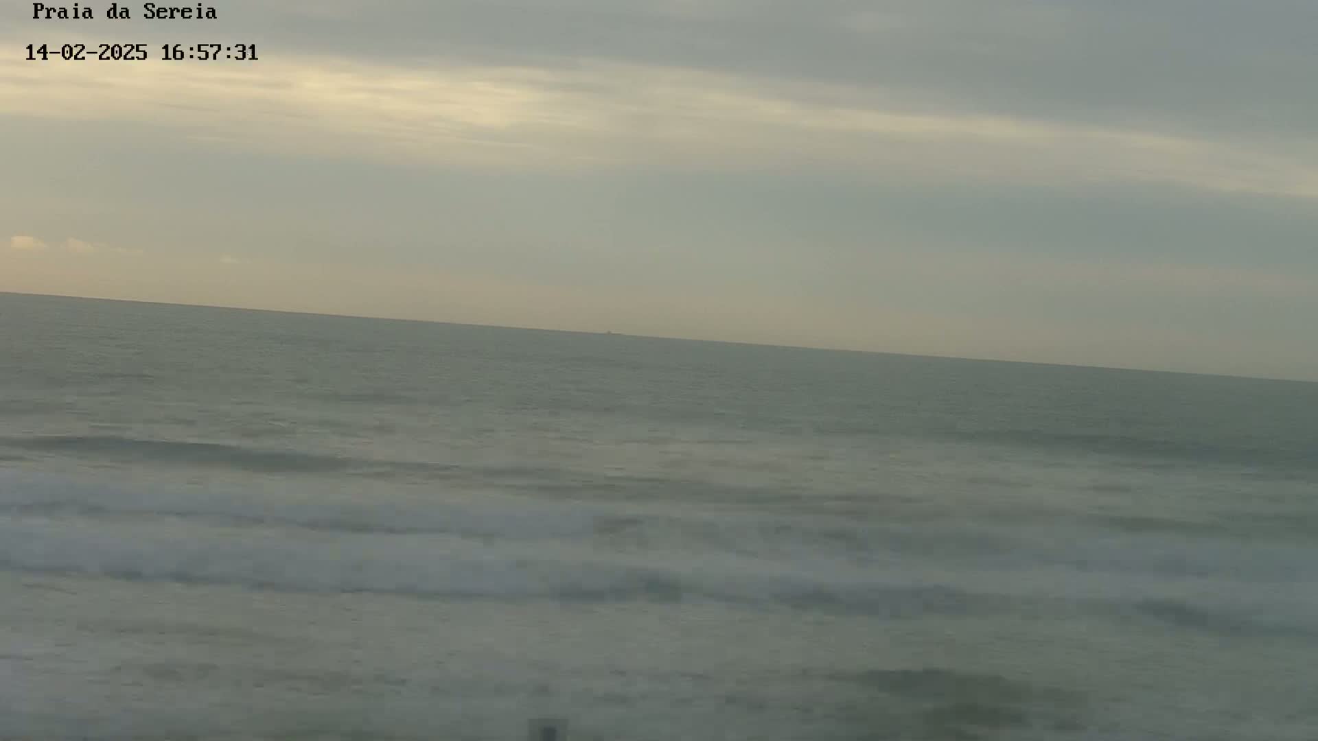 A wide shot of a calm ocean with small waves rolling onto a beach under a cloudy sky.