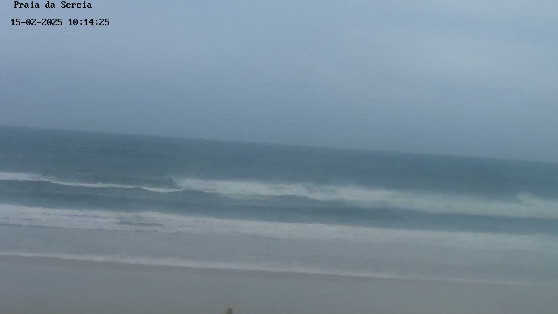 A hazy, overcast day shows a beach with moderate waves rolling onto the shore.