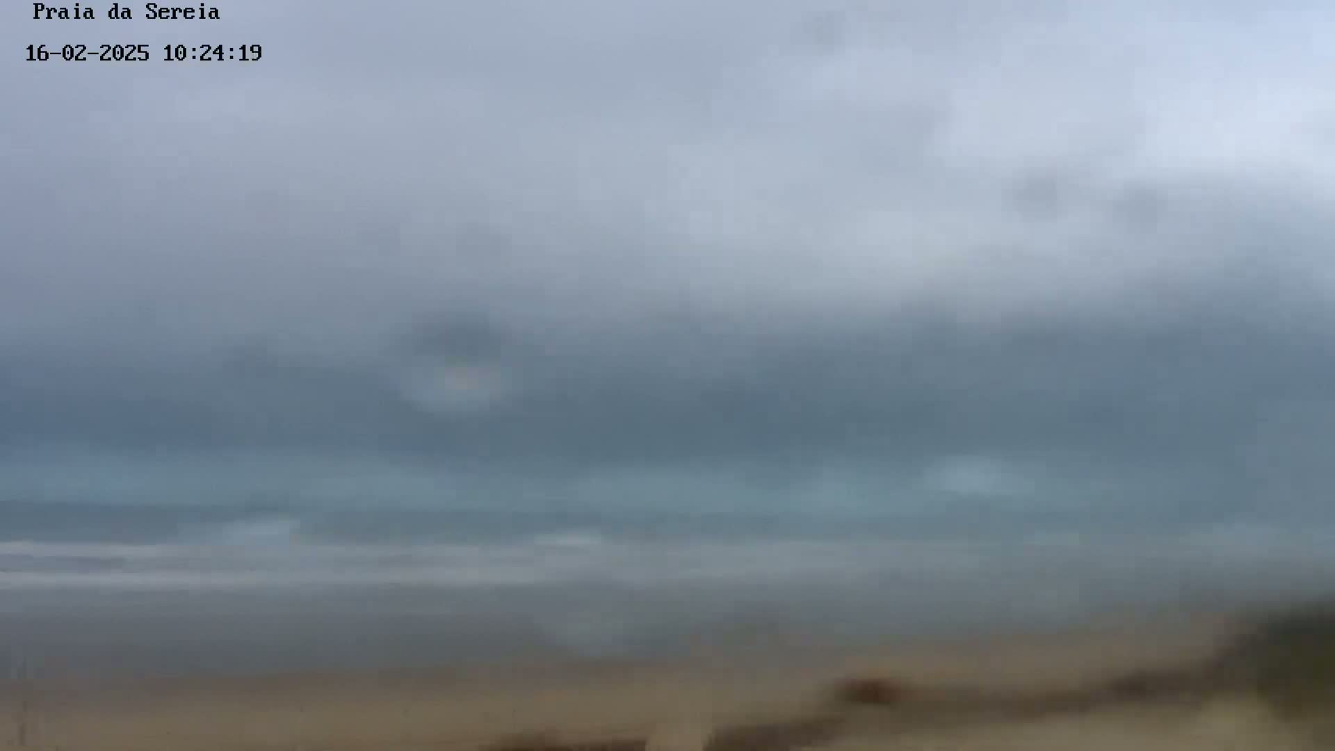 A hazy, overcast sky hangs over a calm ocean and sandy beach.