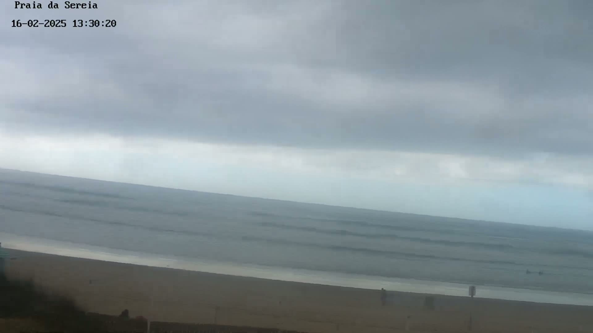 A mostly cloudy sky hangs over a calm ocean with small waves lapping a sandy beach where a few people are walking.