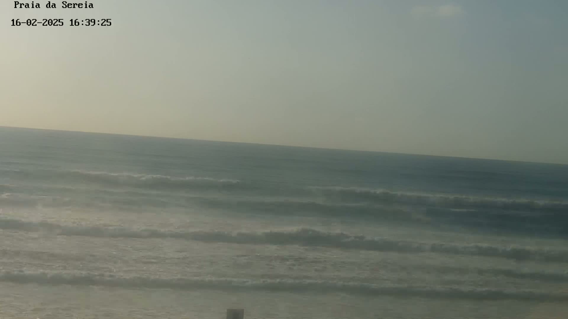 A hazy ocean scene shows moderate waves rolling onto a beach under a light, overcast sky.