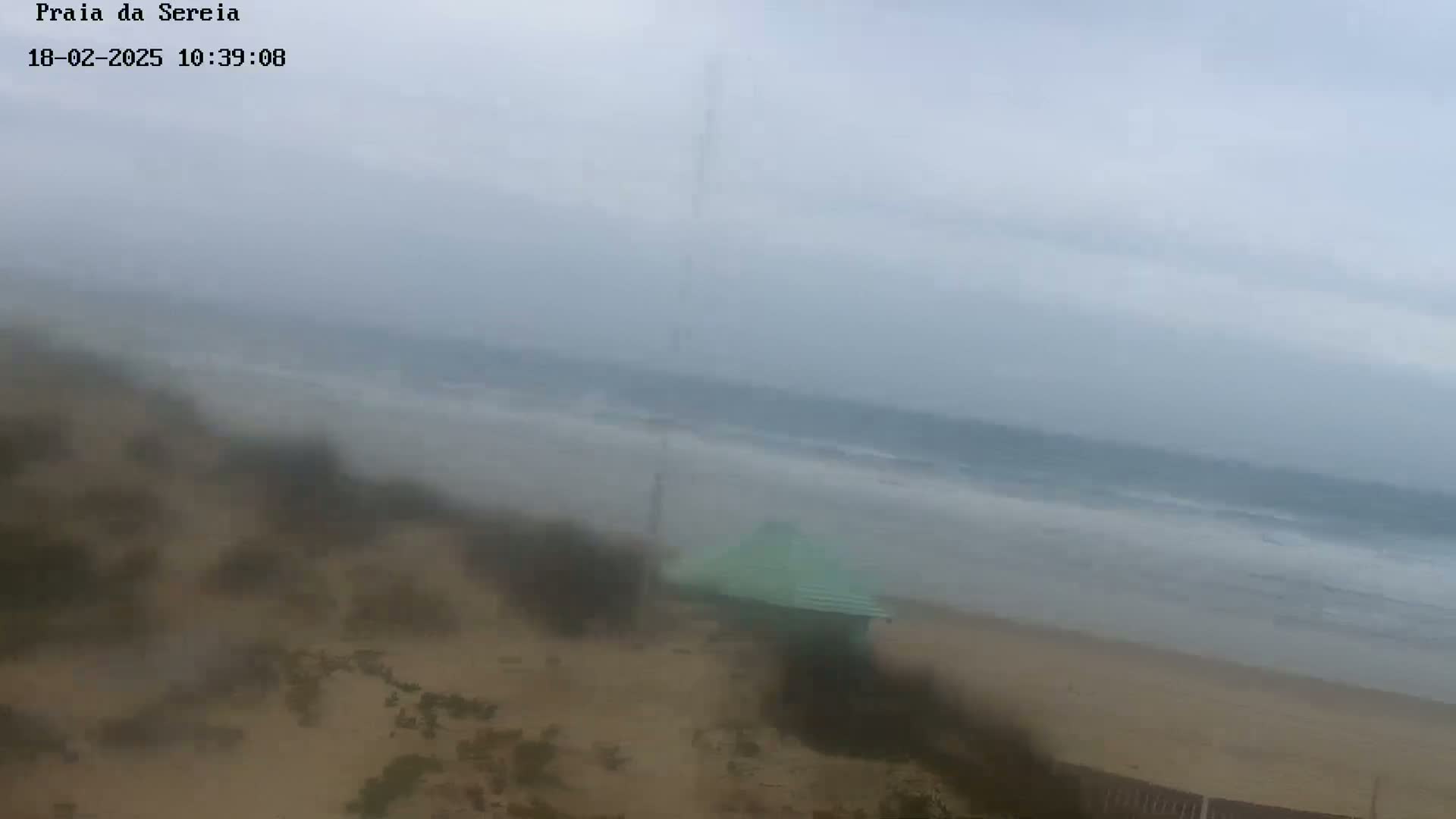 A hazy, overcast day reveals a beach scene with a small, light green structure near the shore and moderate waves breaking in the ocean.