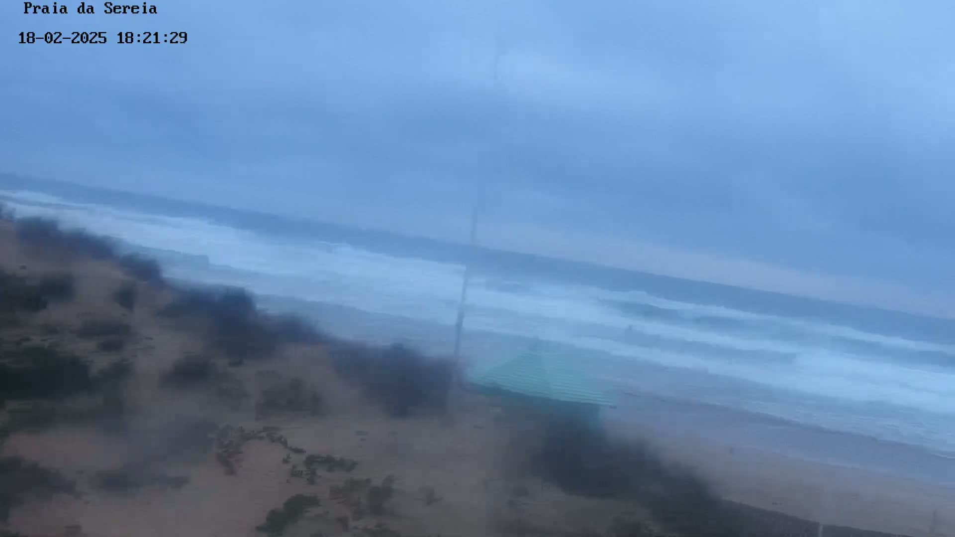 A beach scene shows a somewhat overcast sky, a sandy shore with vegetation, and moderate waves rolling into the ocean.