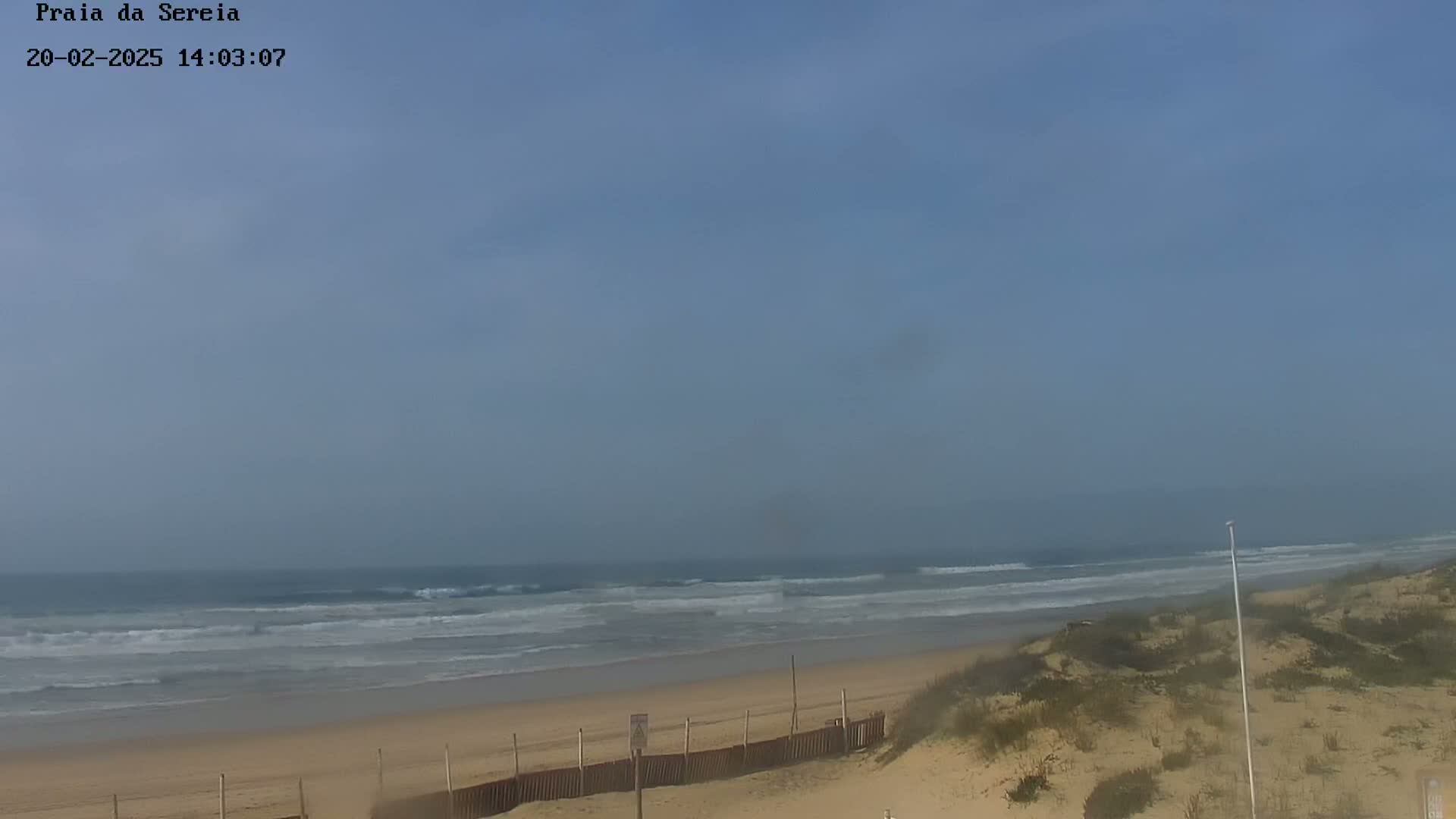 A hazy, sunny day at a beach with gentle waves lapping a sandy shore behind a low dune and fence.