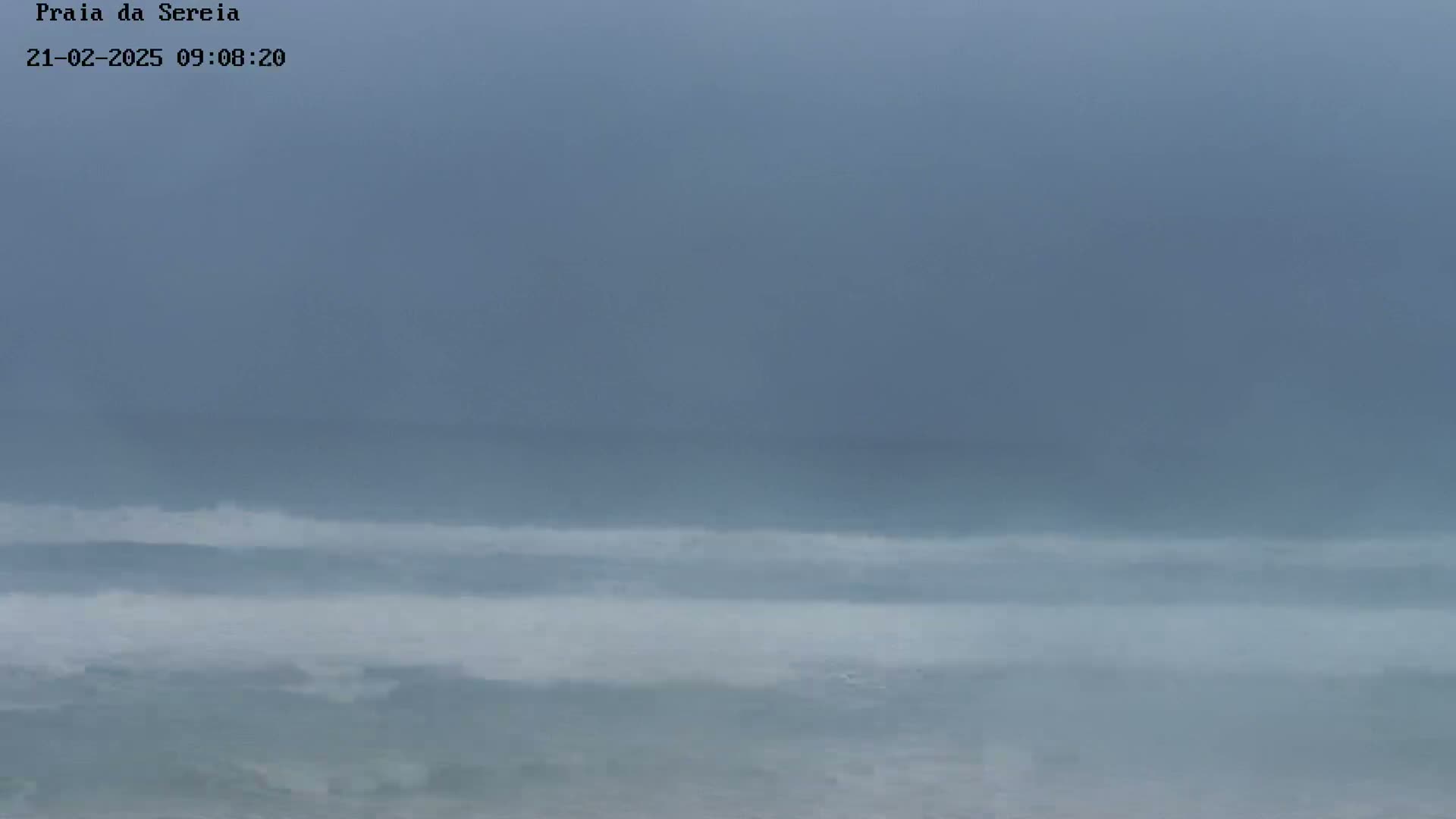 A hazy, overcast sky hangs over a grey ocean with small waves breaking near the shore.