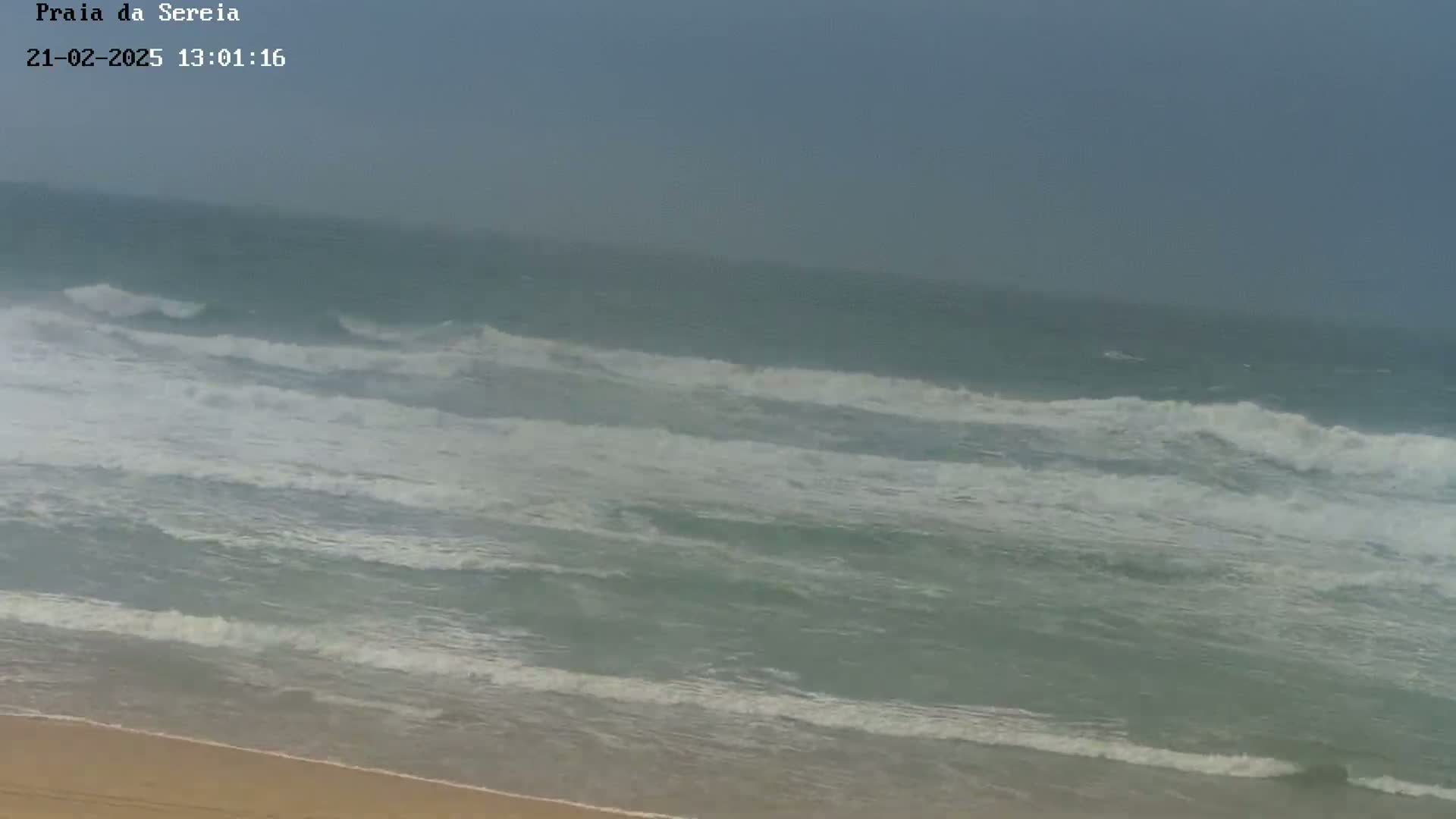 A sandy beach is shown with moderate waves crashing onto the shore under an overcast sky.