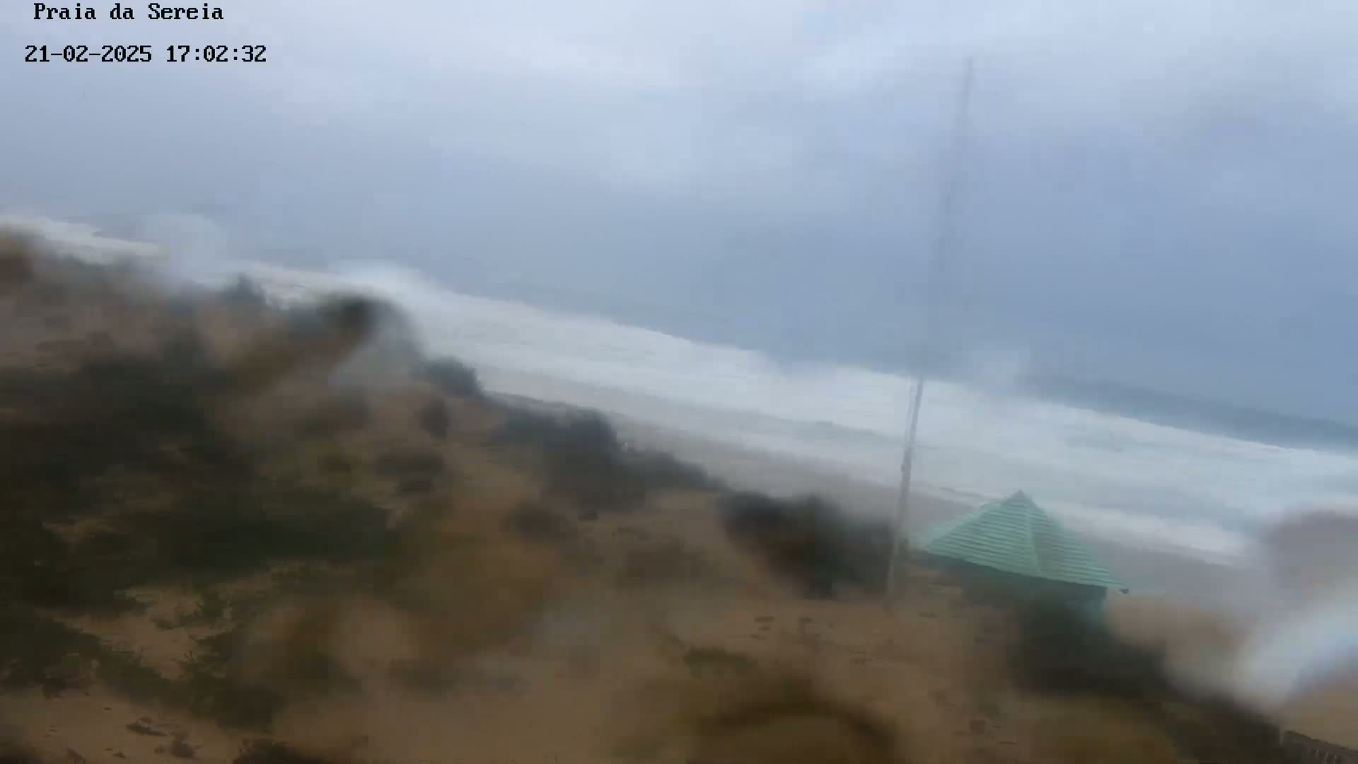 A rain-streaked view shows a beach with turbulent waves under an overcast sky, and a small, pyramid-shaped structure on the sand.