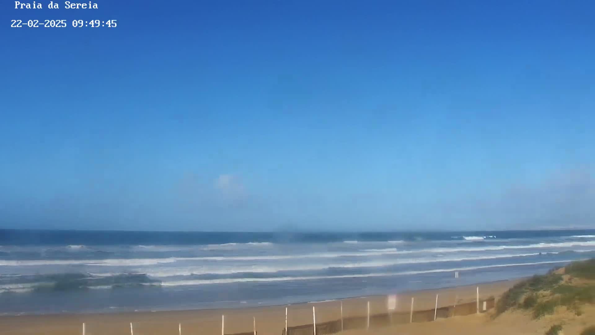 A sandy beach with gentle waves rolls onto the shore under a clear blue sky.