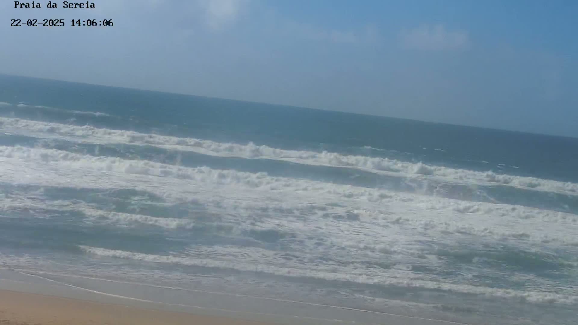 A sandy beach meets a bright blue ocean with moderate waves under a mostly clear sky.