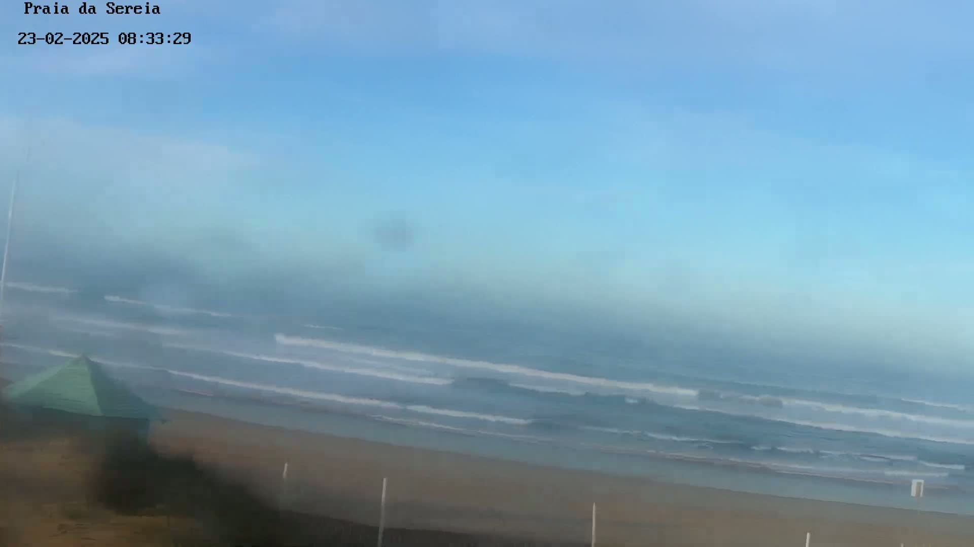 A hazy, mostly clear sky overlooks a sandy beach with gentle waves rolling in on an ocean.