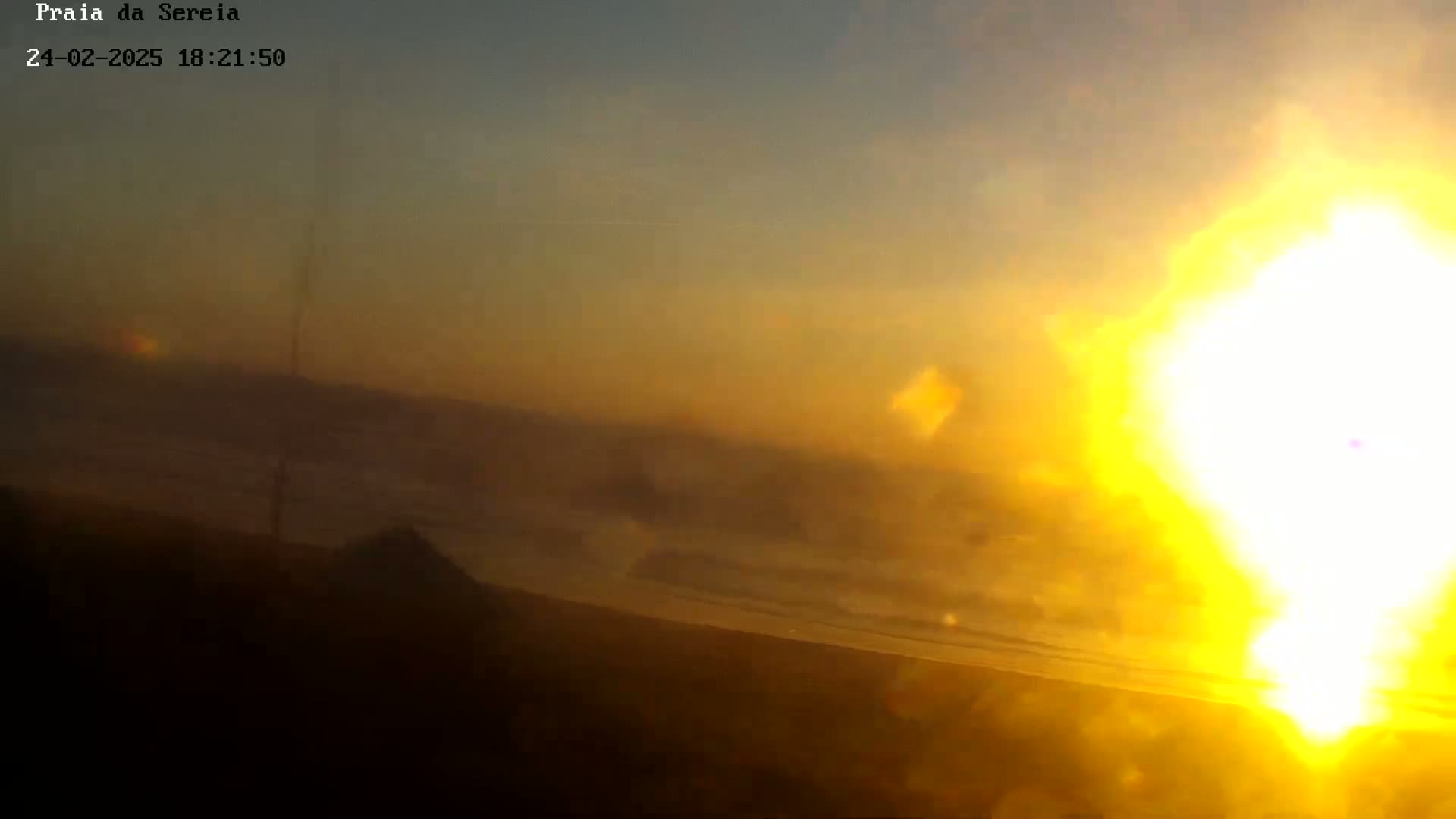 A hazy, golden sunset over a dark landmass, possibly a beach, with the sun appearing bright and blurry.