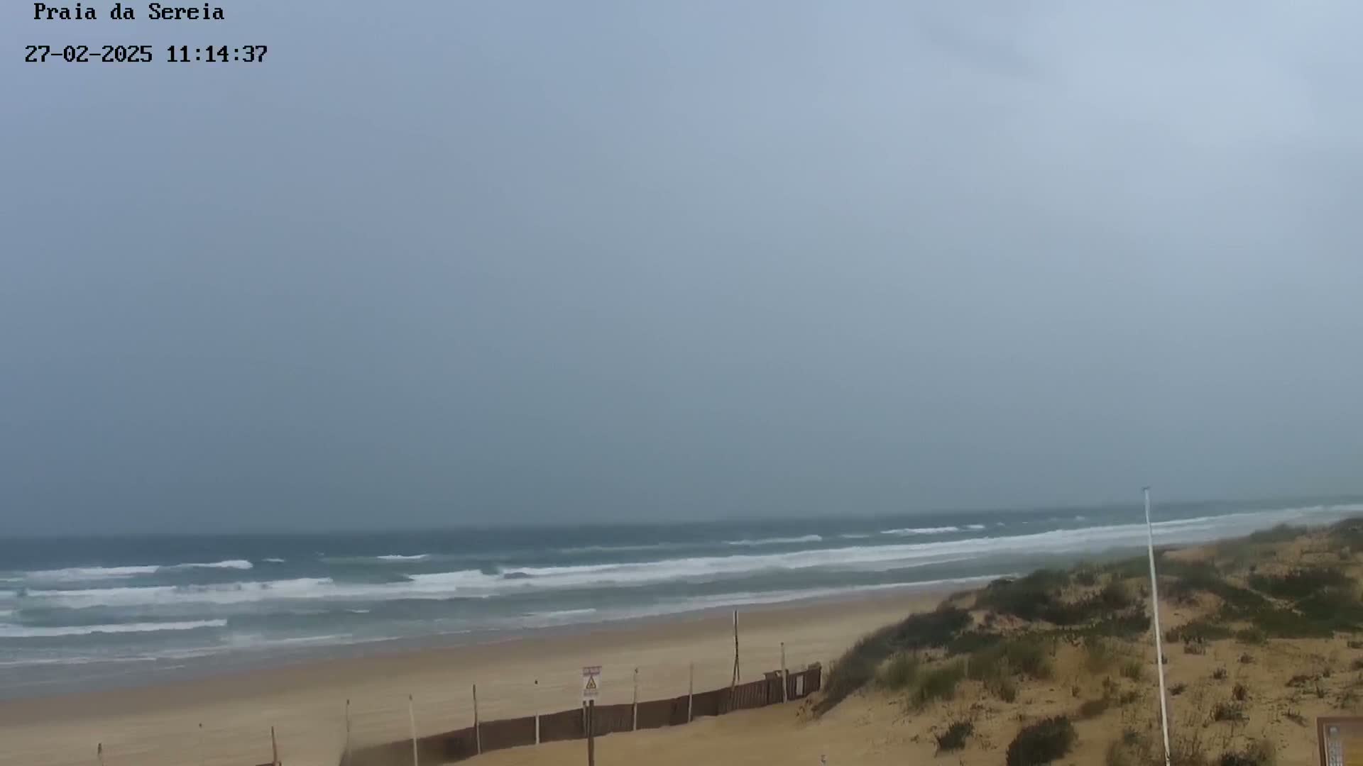 A sandy beach with moderate waves under an overcast sky is seen from a dune.