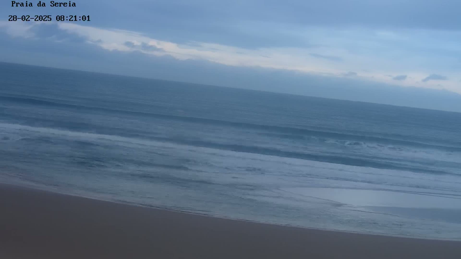 A sandy beach slopes down to a calm ocean under a mostly cloudy sky.