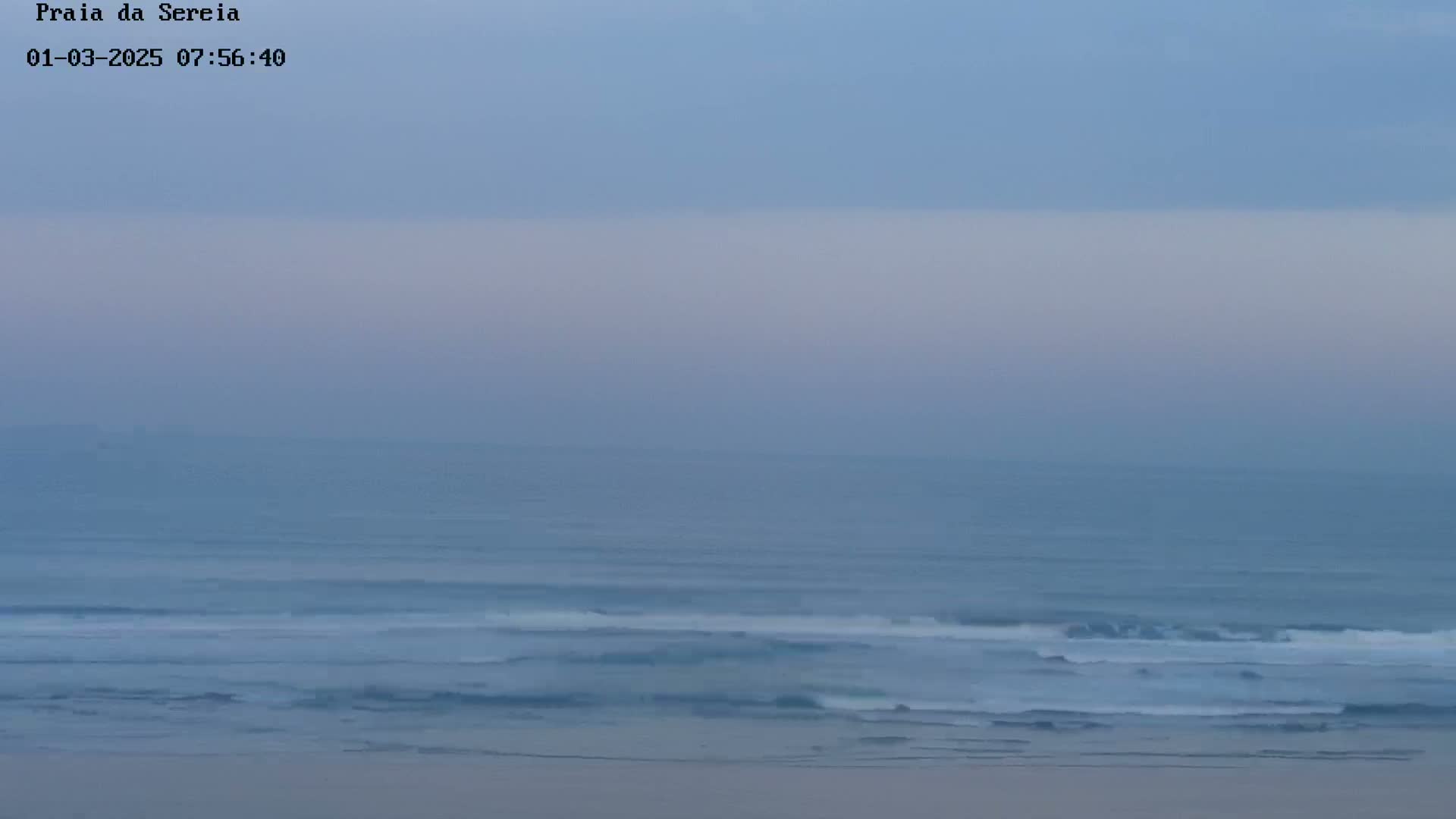 A hazy, light-blue sky overlooks a calm ocean with small waves breaking on the shore.