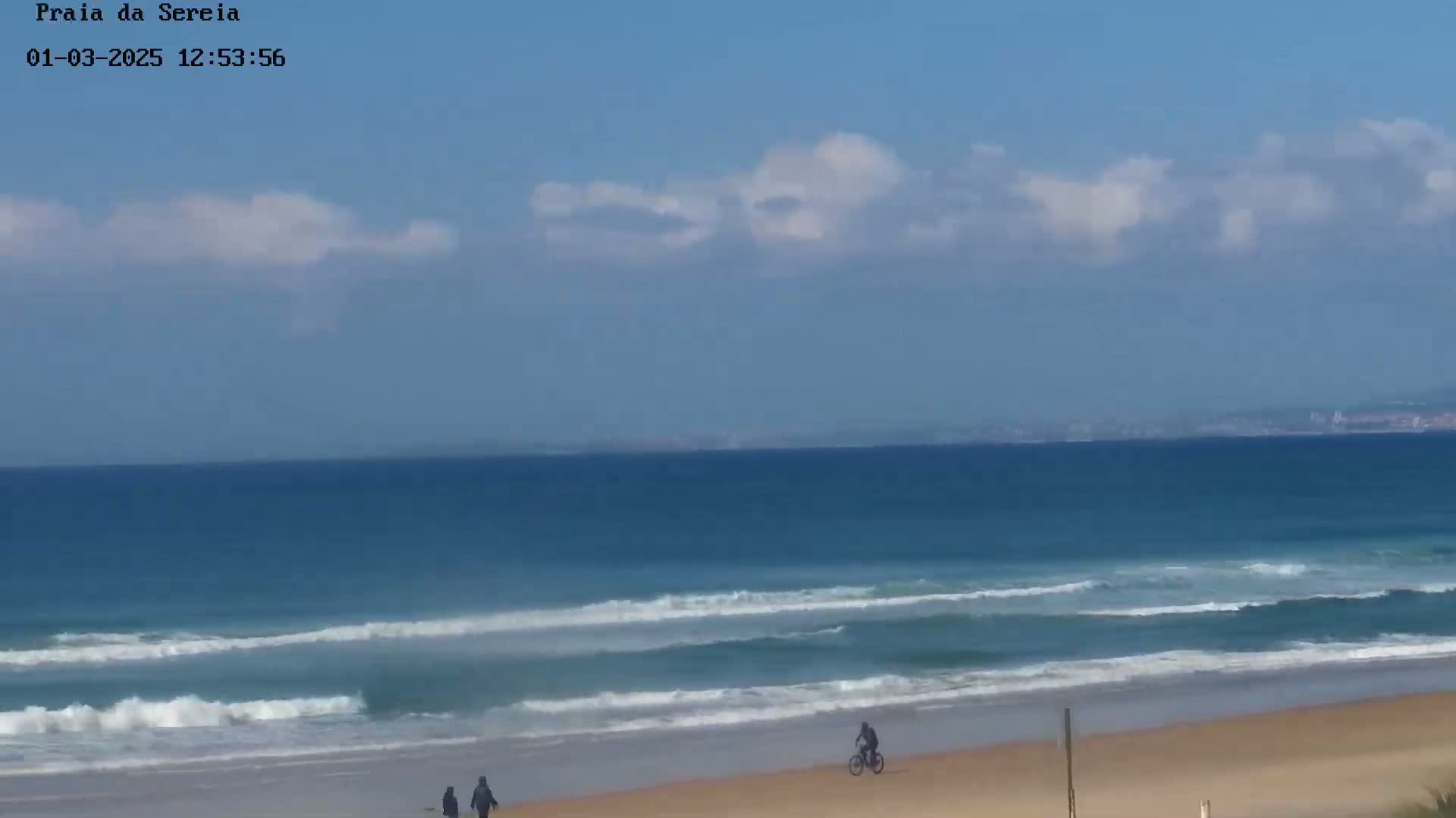 A mostly sunny day at a beach shows a calm ocean with small waves lapping a sandy shore where a few people stroll and a person rides a bicycle.