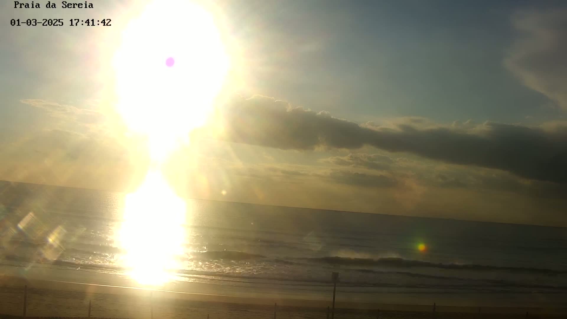 A bright, sun-filled sunrise over a calm ocean with a few clouds in the sky and a sandy beach in the foreground.