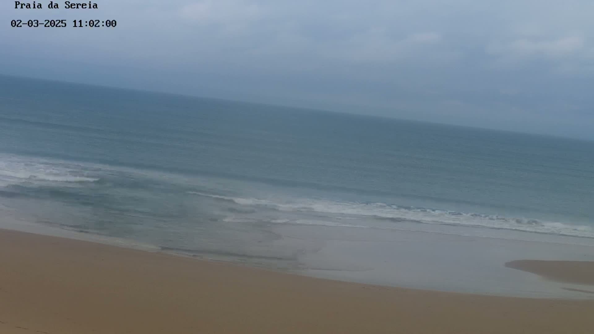 A sandy beach slopes down to a calm, gray-blue ocean under a cloudy sky.