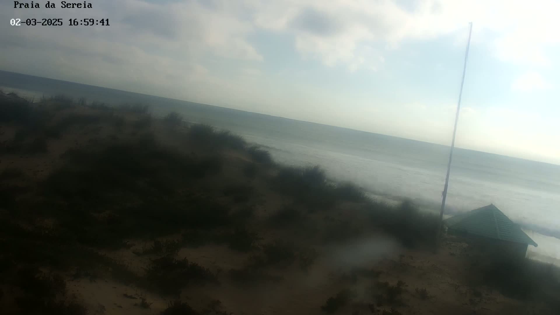 A hazy view of a beach with a small green building and a tall pole, under an overcast sky.