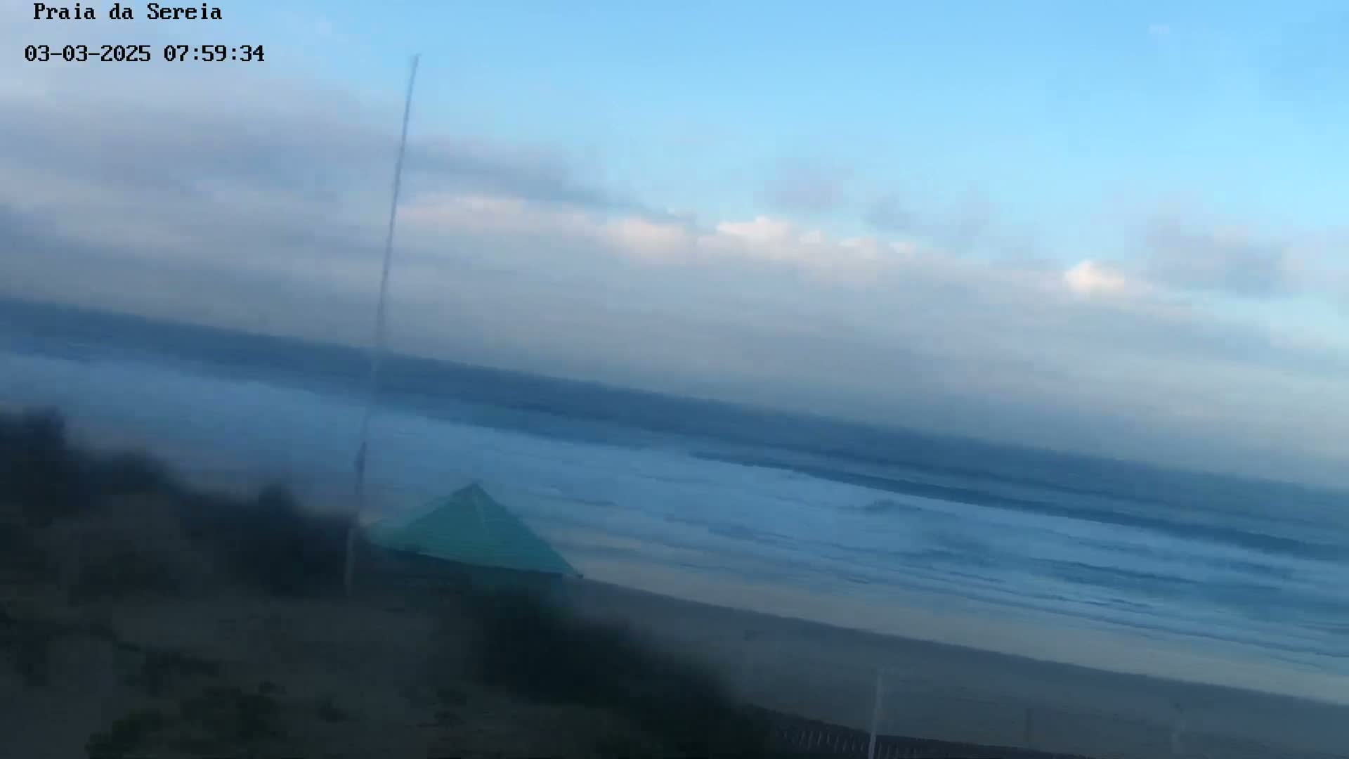 A mostly cloudy sky overlooks a beach with a small, teal-colored structure and a somewhat choppy ocean.