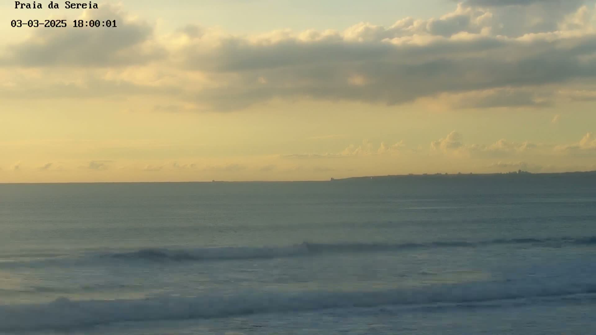 A calm ocean with small waves under a partly cloudy, yellow-tinged sky shows a distant coastline.