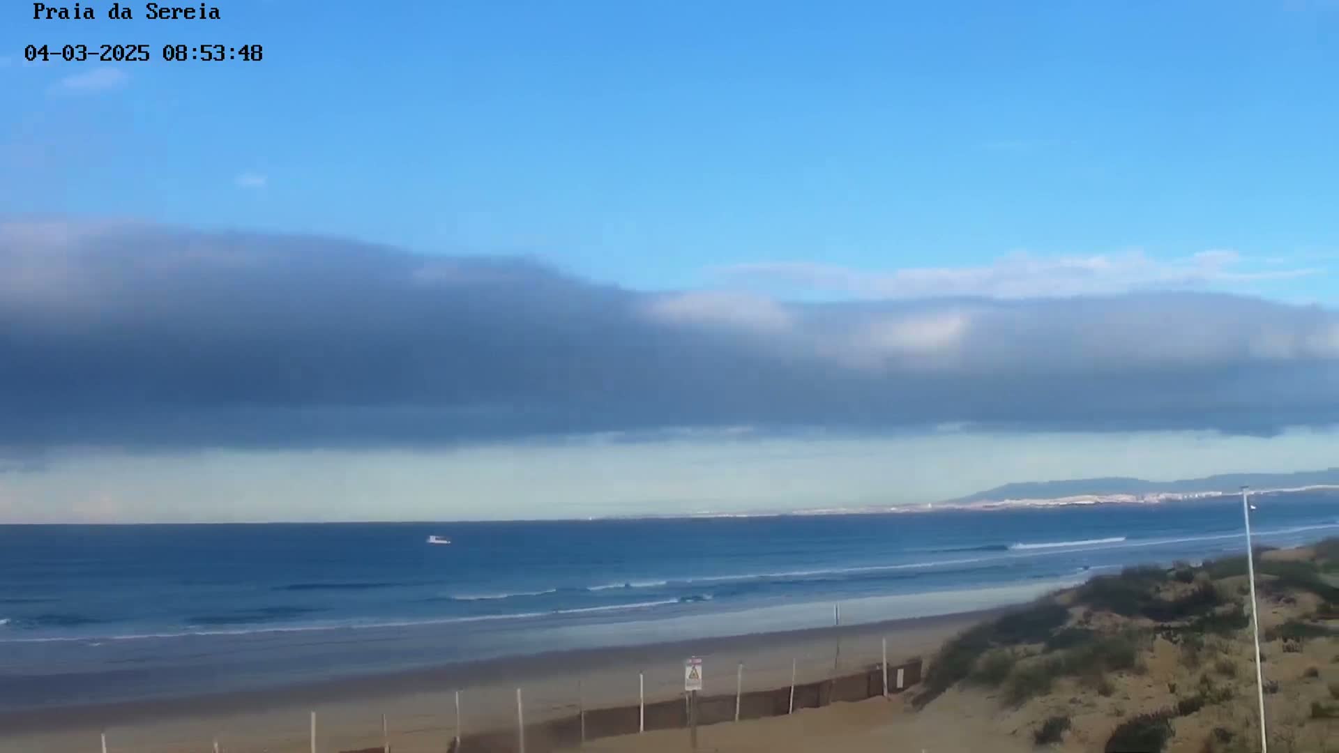 A mostly sunny beach scene shows a long stretch of ocean with small waves under a layer of low-lying clouds.