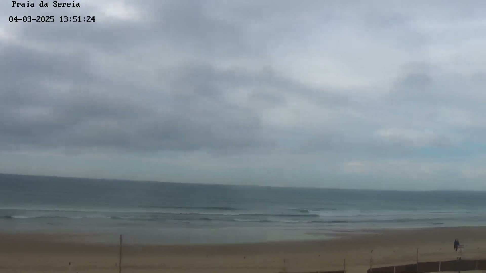 A mostly cloudy sky overlooks a calm ocean with small waves lapping a sandy beach.
