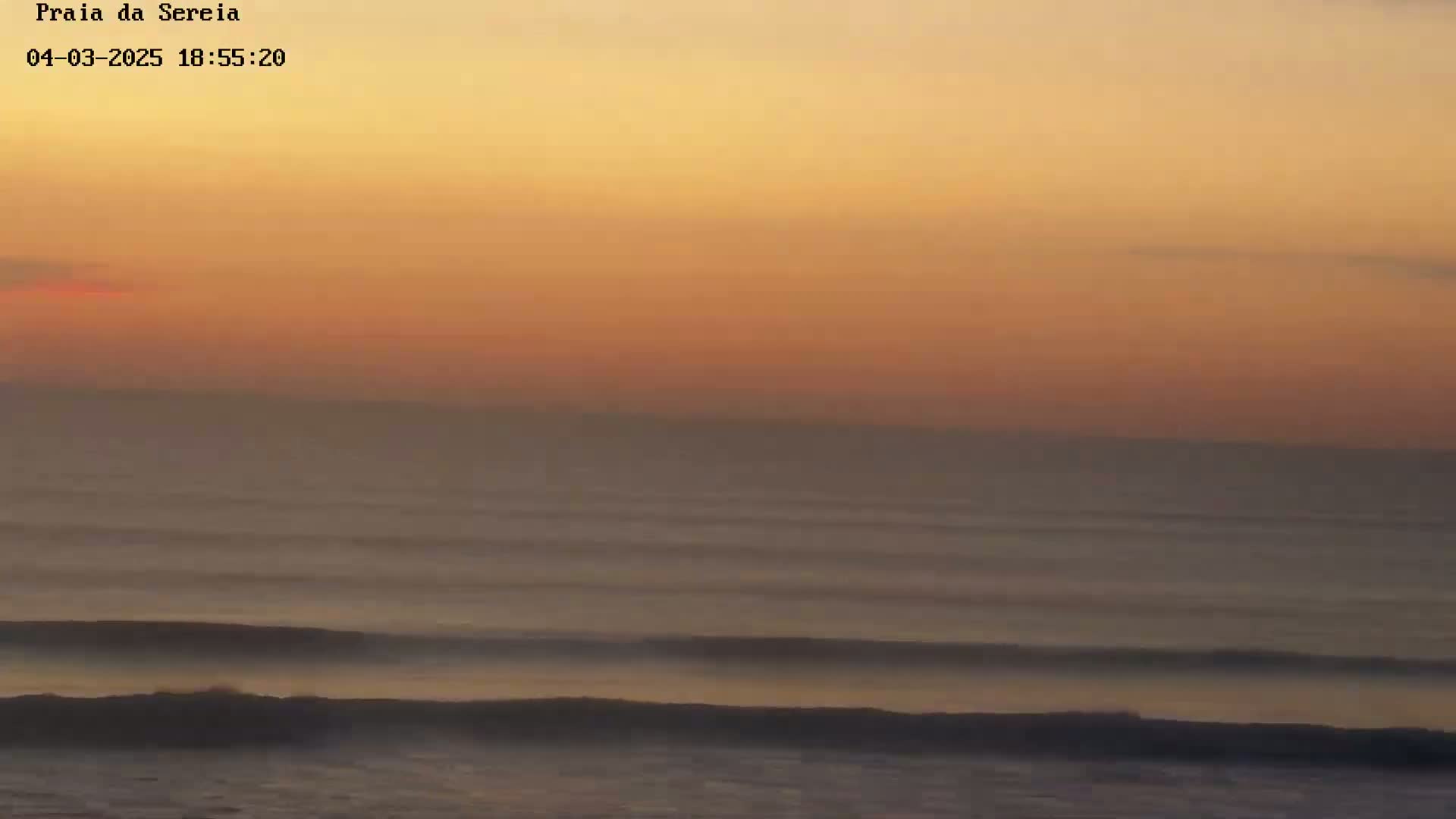 Gentle waves roll onto a hazy beach under a warm, orange-hued sunset sky.