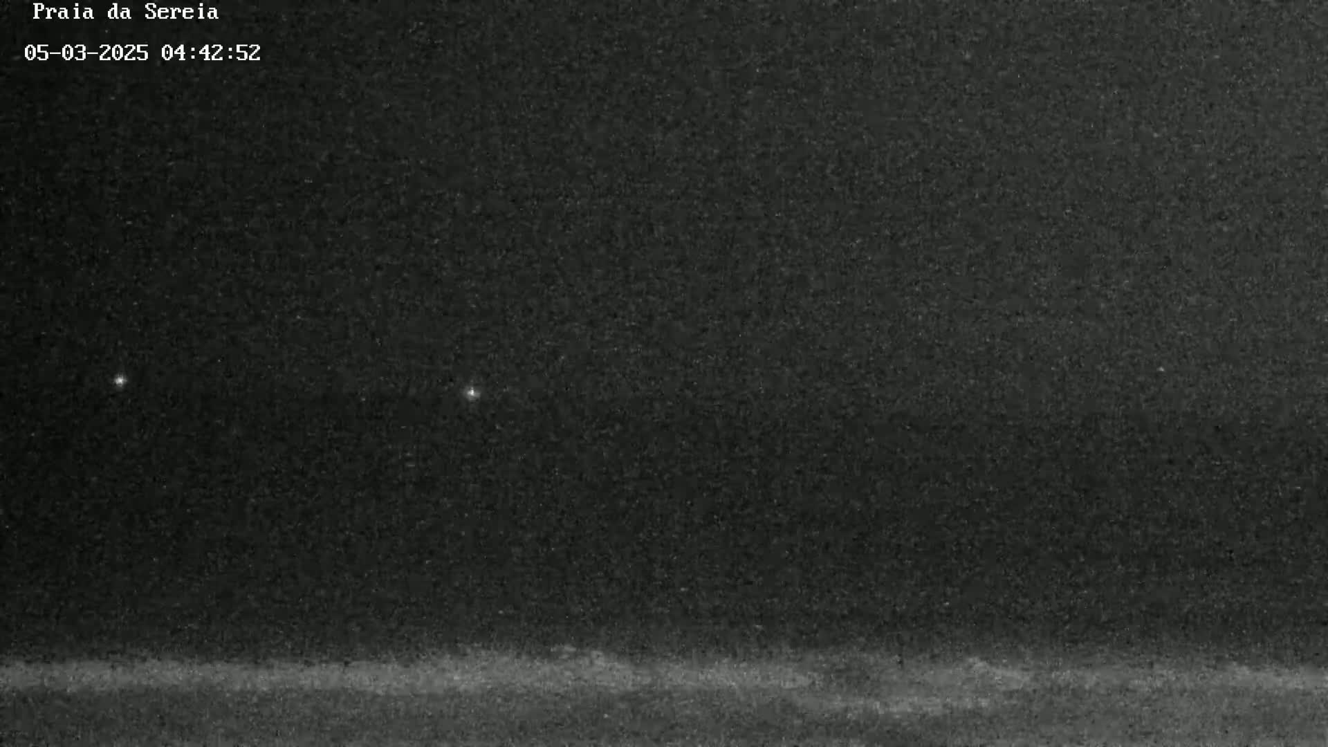A nighttime, low-light image shows a dark, possibly sandy landscape under a mostly clear, dark sky with two faint lights visible in the distance.