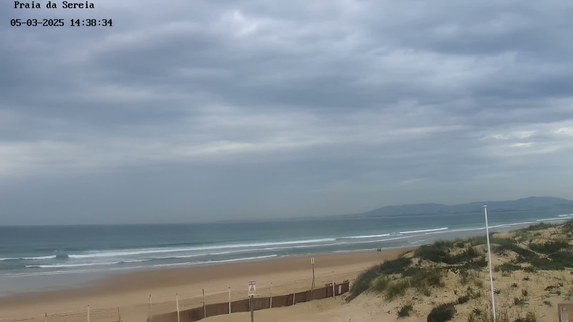 A sandy beach with gentle waves rolls into a calm ocean under a cloudy sky, with distant hills visible on the horizon.