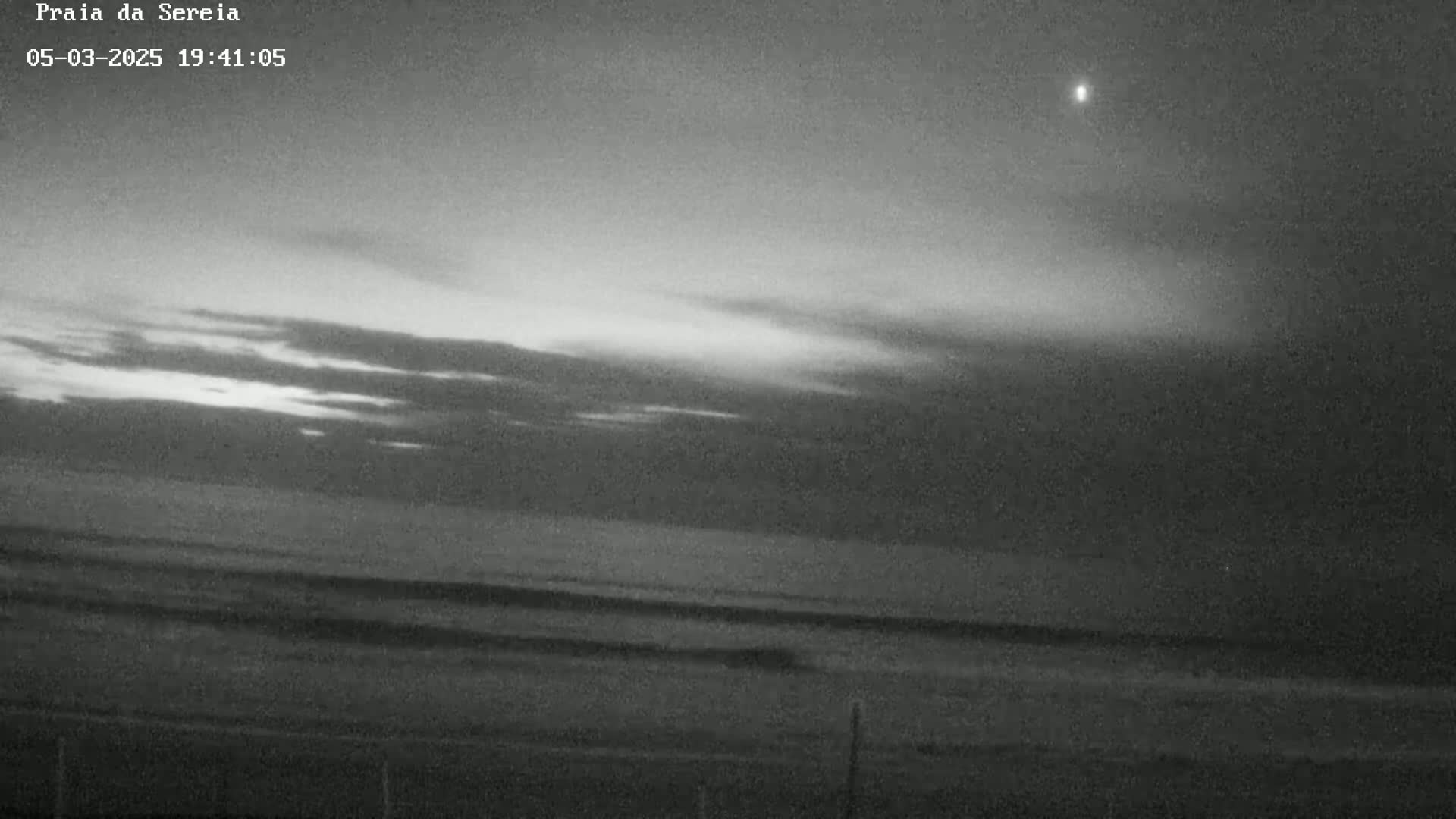 A grayscale image shows a dark, overcast sky above a calm ocean meeting a dark beach at dusk.