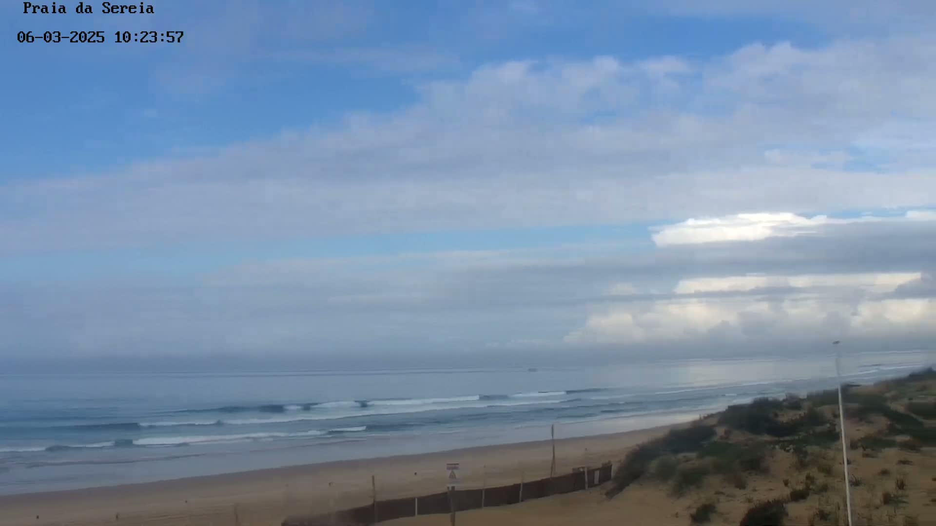 A partly cloudy sky overlooks a sandy beach with gentle waves rolling into shore.