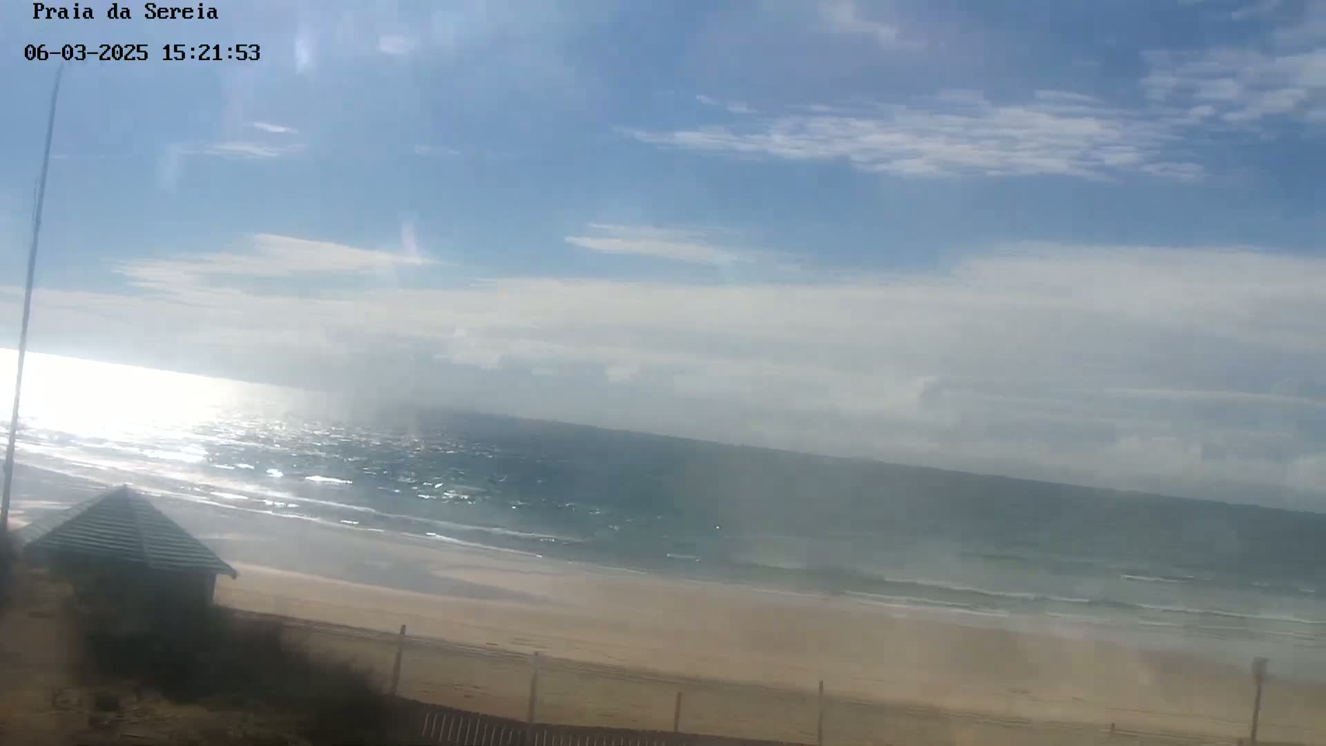 A sandy beach with a small building and a partly cloudy sky overlooks a wavy ocean under sunny conditions.