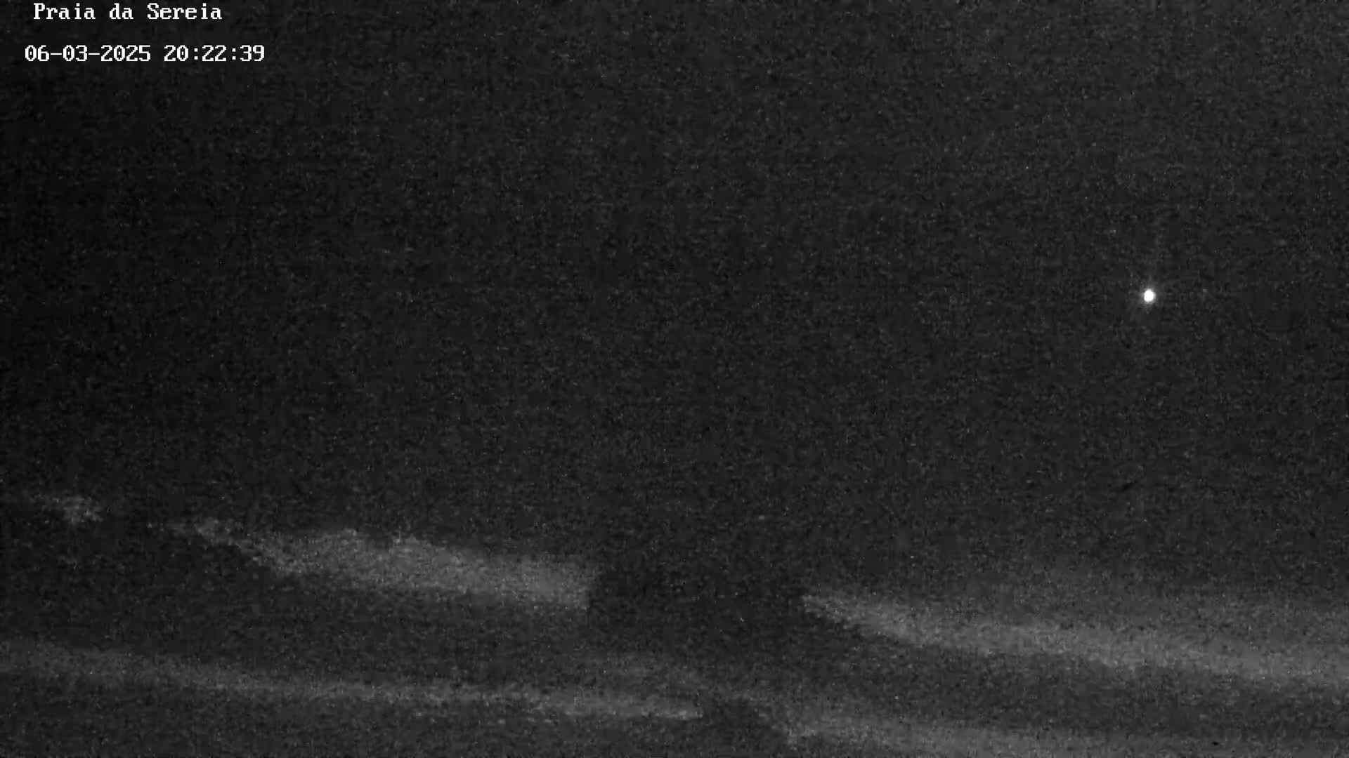 A nighttime, low-light image shows dark, wavy terrain under a mostly clear sky with a single bright light visible.