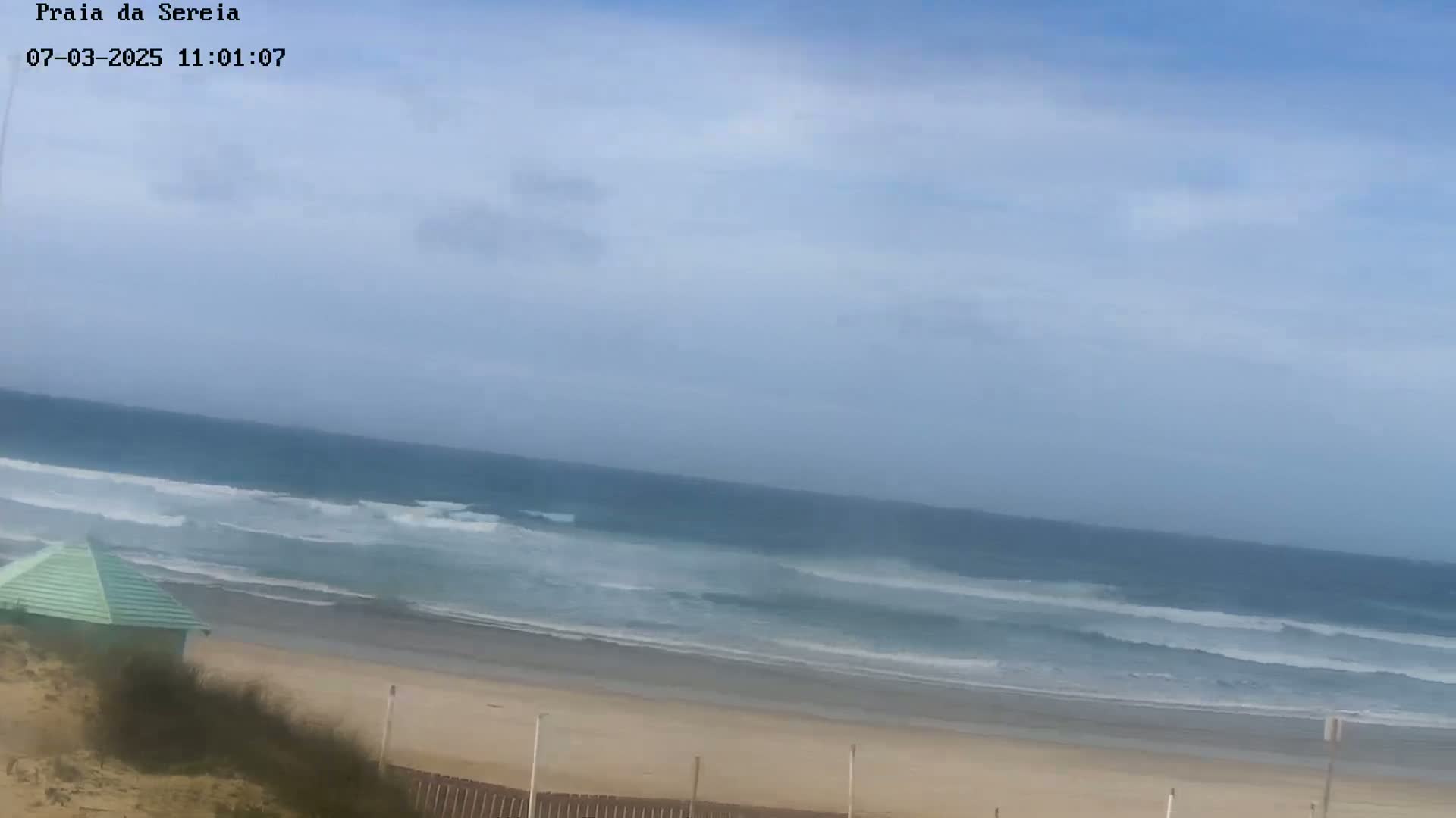 A mostly sunny day at a sandy beach shows a calm ocean with small waves, a small light-green beach hut, and a wooden fence.