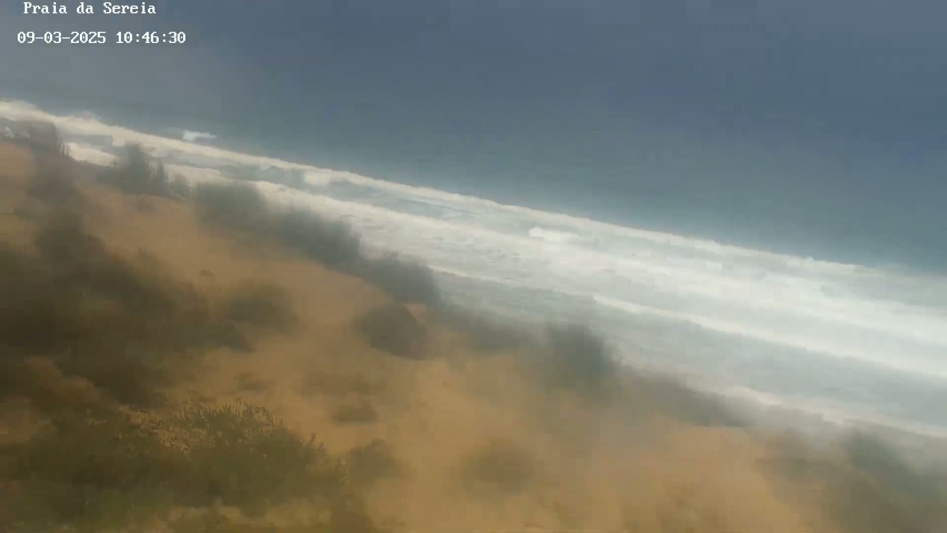 A hazy view shows a sandy dune overlooking a beach with moderately sized waves under a mostly cloudy sky.