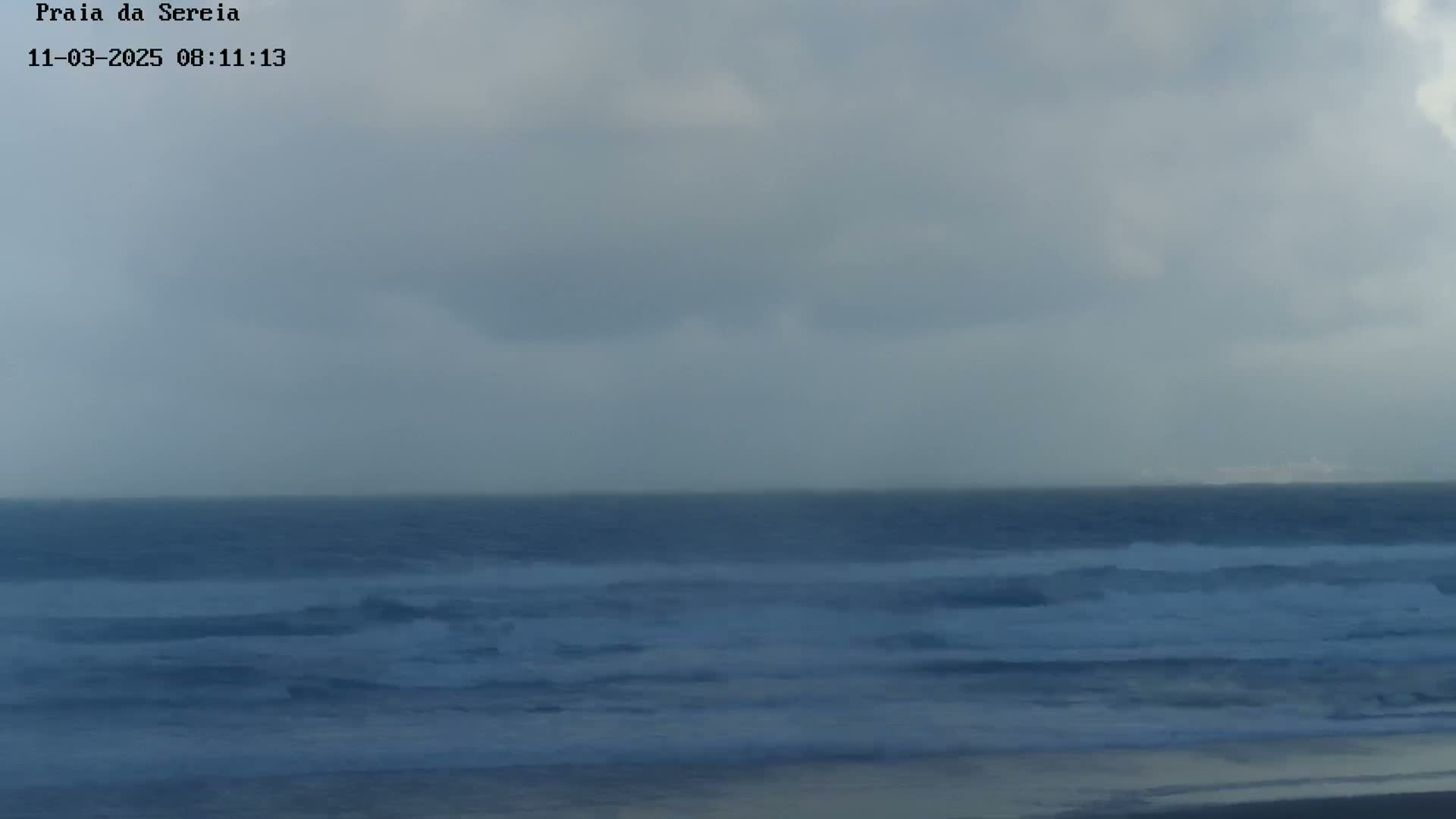 A dark, overcast sky hangs over a restless ocean with small waves breaking on a sandy beach.