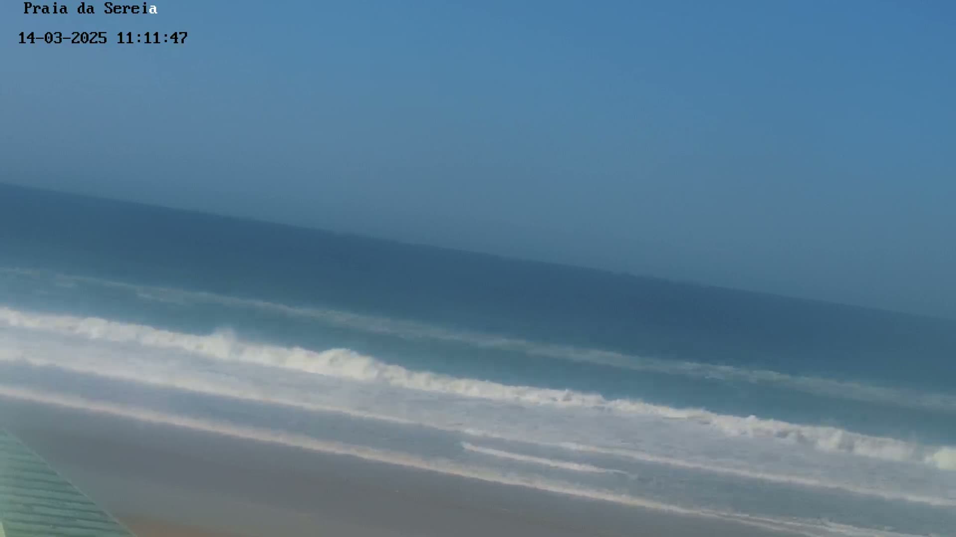 A sandy beach meets a blue ocean with moderate waves under a clear sky.