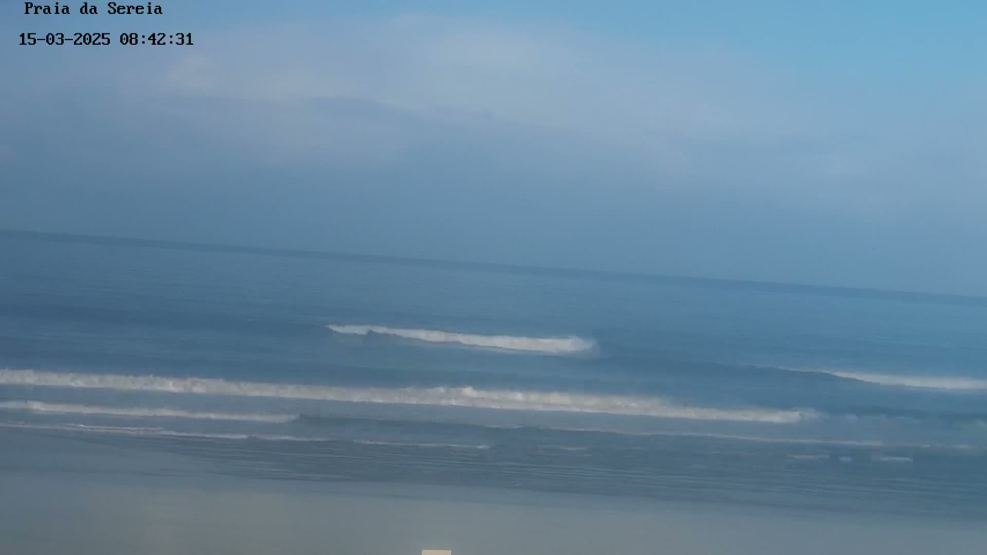 A hazy, calm ocean with small waves rolls onto a mostly obscured beach under a clear, light blue sky.