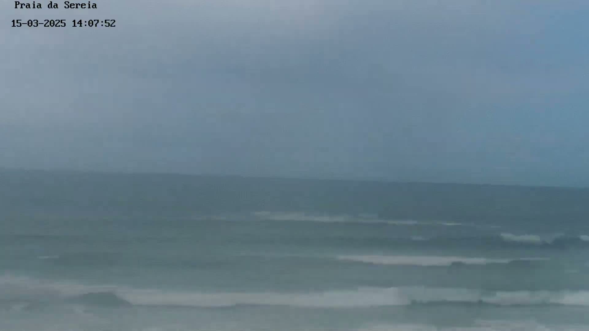 A hazy, overcast sky overlooks a choppy ocean with small waves breaking on the shore.