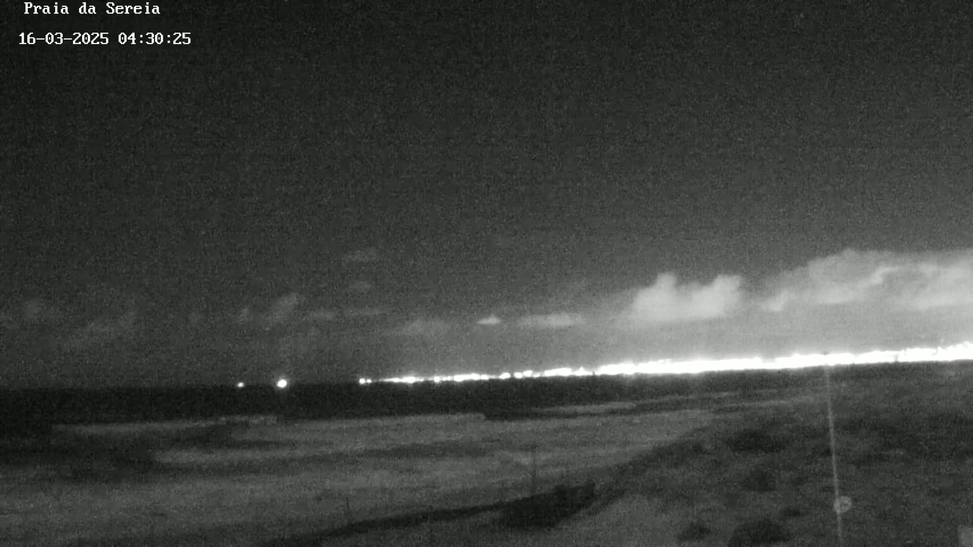A nighttime, low-light view of a dark beach with a distant line of lights on the horizon under a mostly cloudy sky.