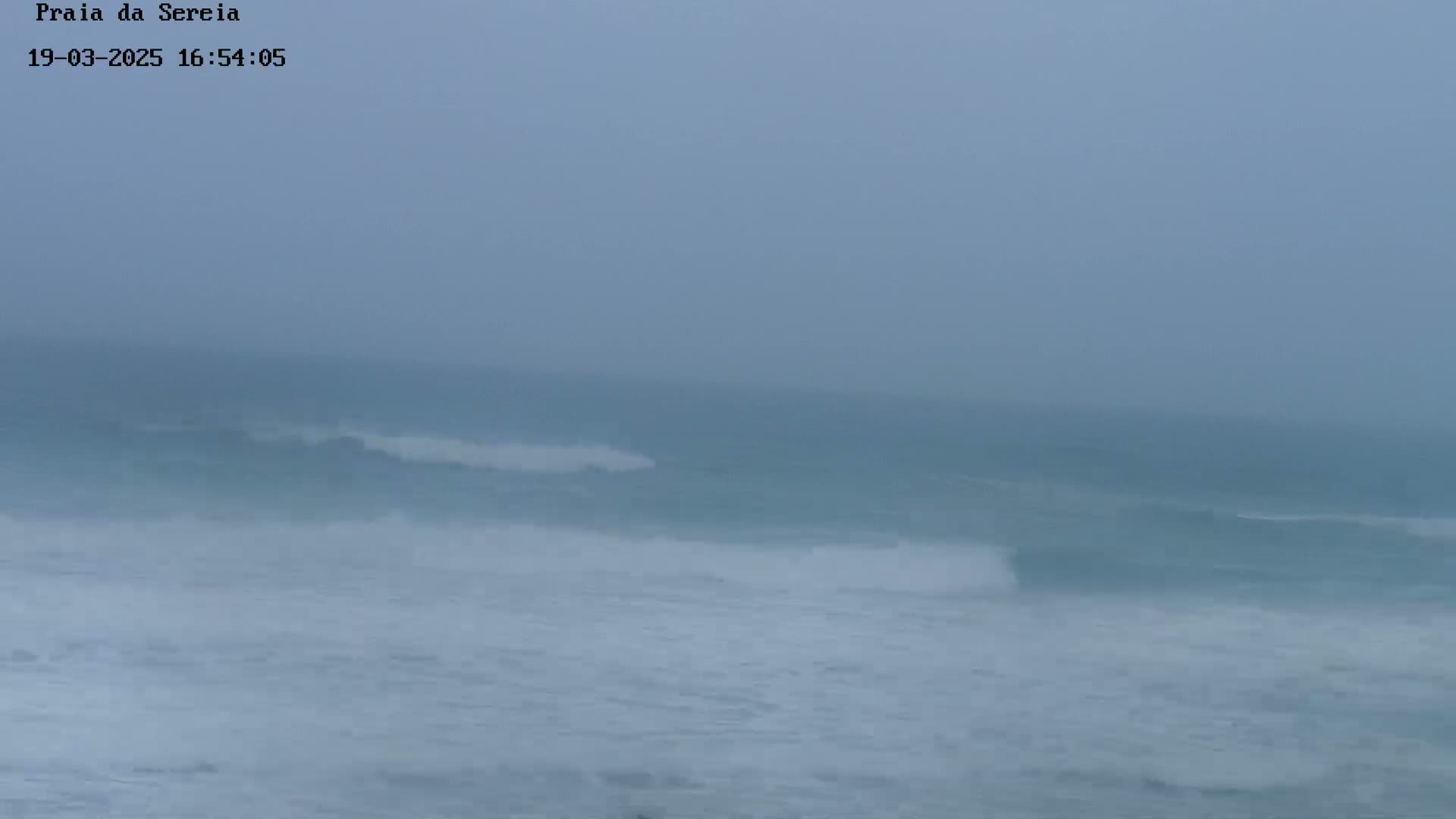A hazy, overcast day shows moderate waves rolling into a shore.
