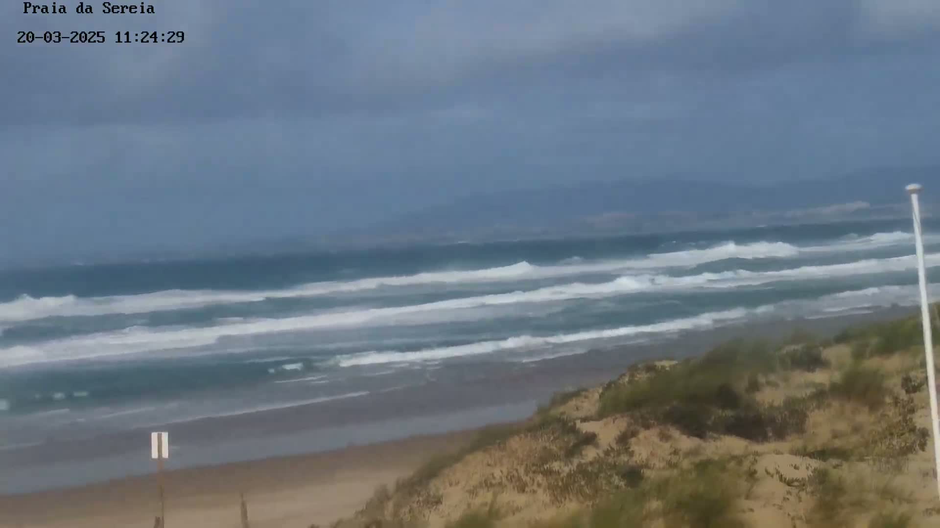 Under a mostly cloudy sky, moderate waves roll onto a sandy beach backed by dunes with sparse vegetation.