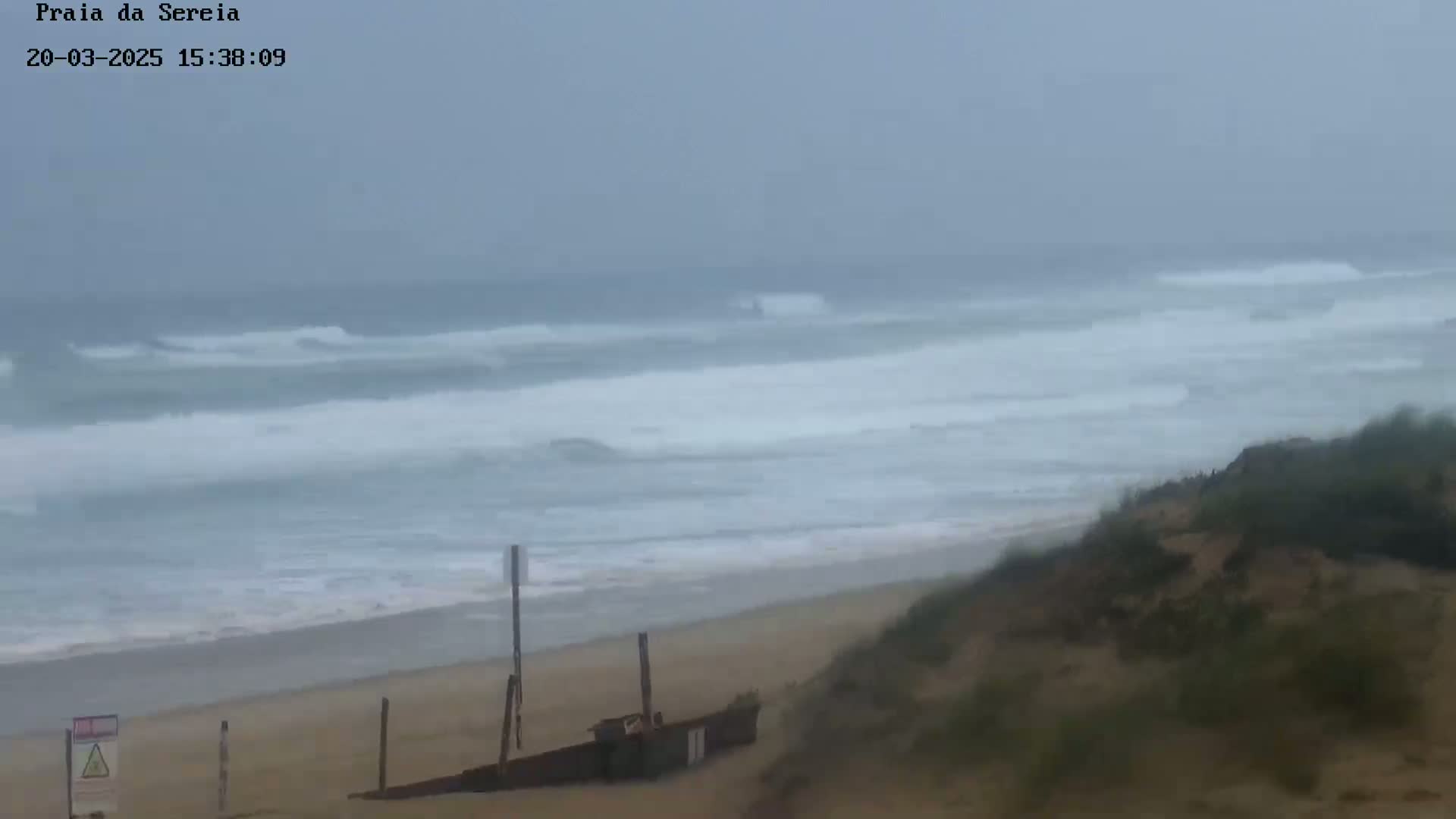 A hazy, overcast day shows a beach with moderate waves rolling onto the shore, partially obscured by a sand dune with sparse vegetation.