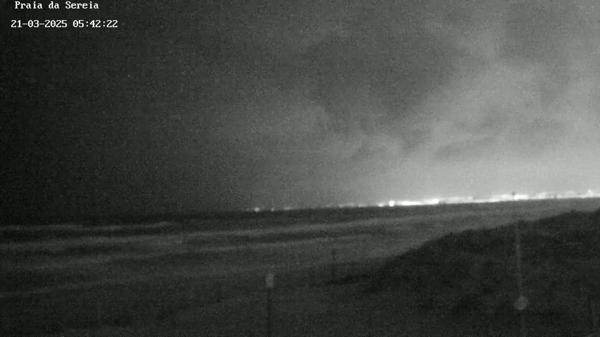 A dark, nighttime beach scene shows a dark ocean under a cloudy sky with distant lights on the horizon.