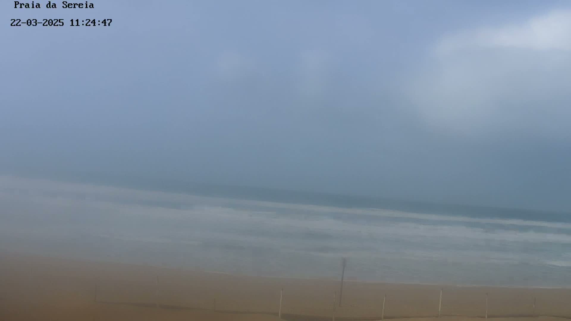 A hazy, foggy day at a beach shows a sandy shore and ocean waves under a mostly cloudy sky.